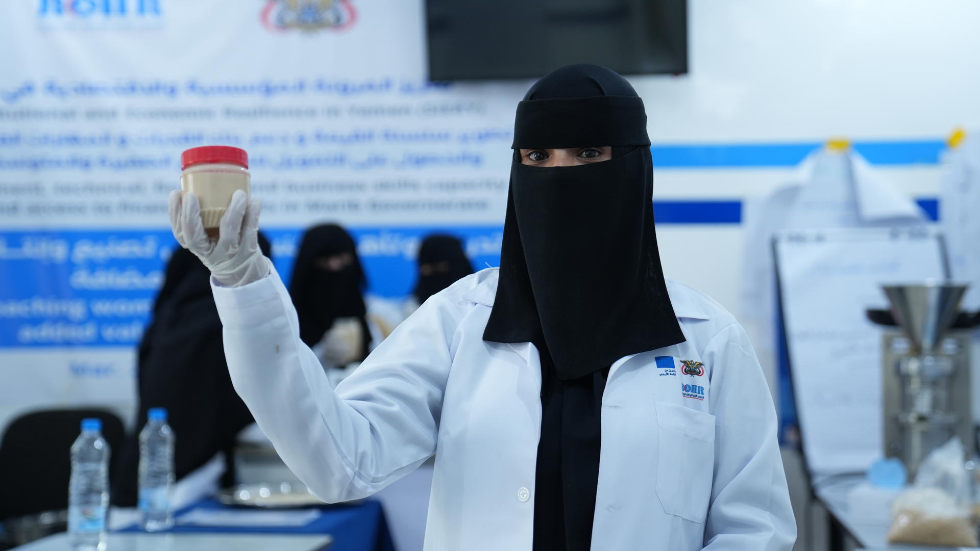 A woman in a lab coat and niqab holds a jar, surrounded by a professional setting.