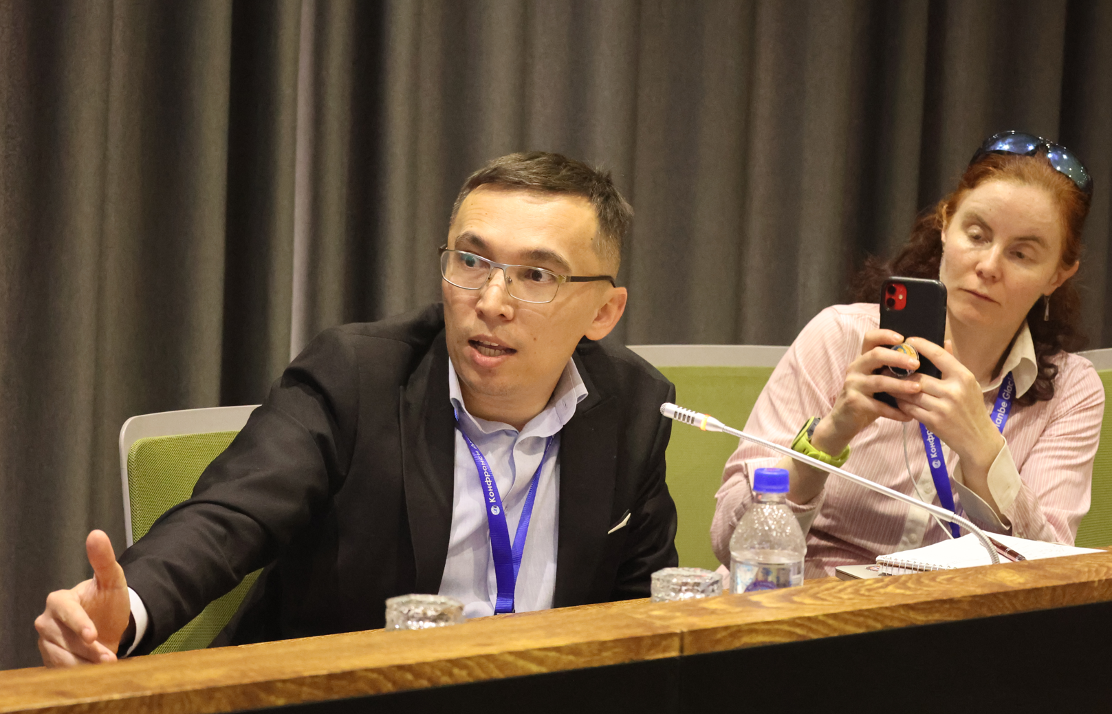 A man in a suit gestures while speaking; a woman in the background takes notes on her phone.