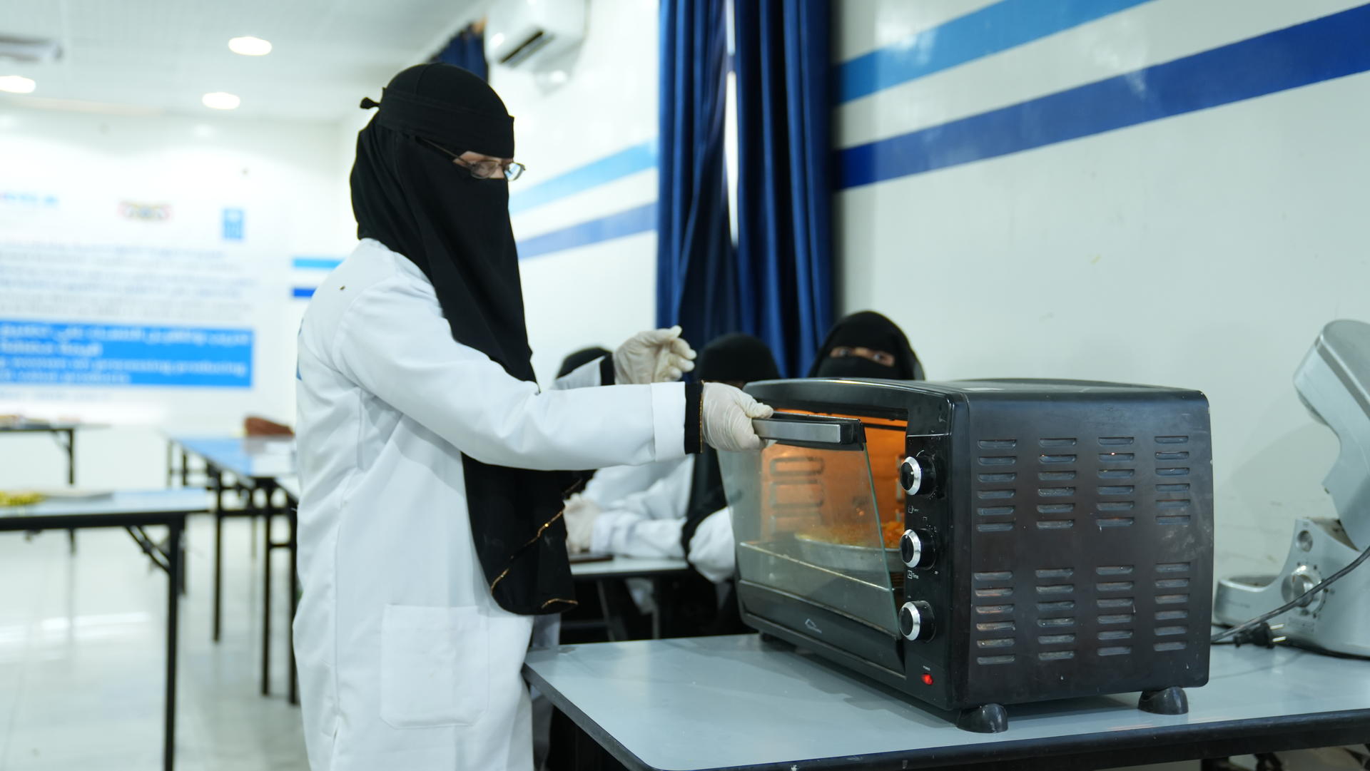 A person in a lab coat and niqab operates a toaster oven in a bright classroom setting.
