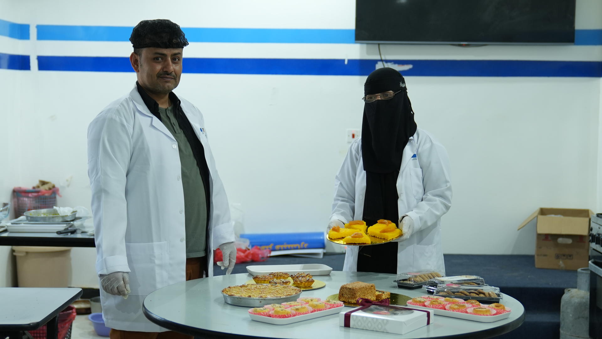 A man and a woman in lab coats stand by a table with various food items.