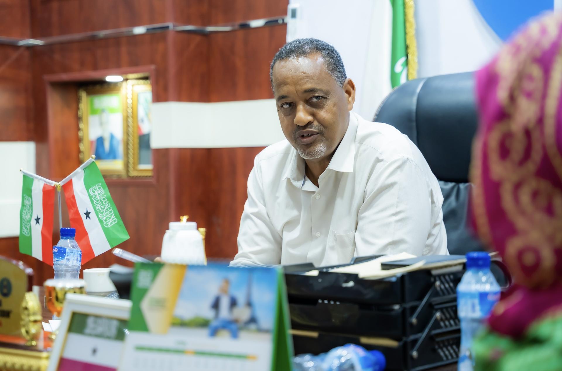 A man in a white shirt sits at a desk, engaging with someone out of view. Flags are visible.