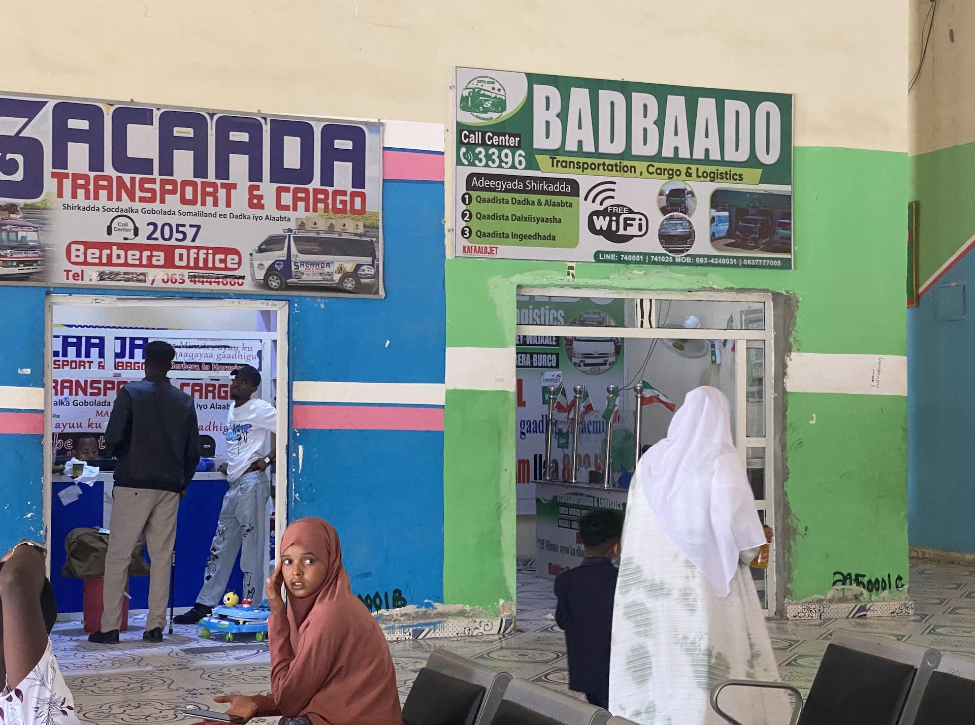 A busy transport office with two shops, one labeled "Acaada" and the other "Badbaado," and people inside.