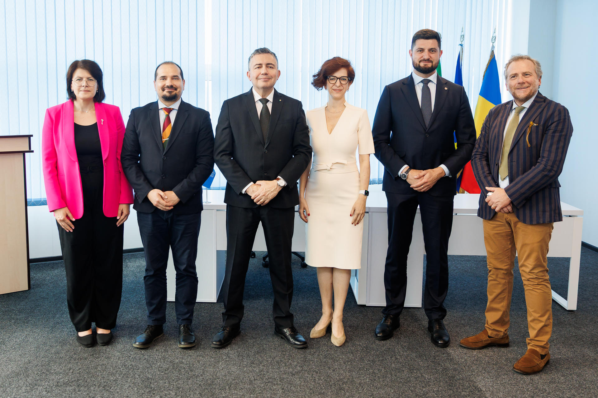 A group of six professionals poses together in formal attire, smiling, with flags in the background.