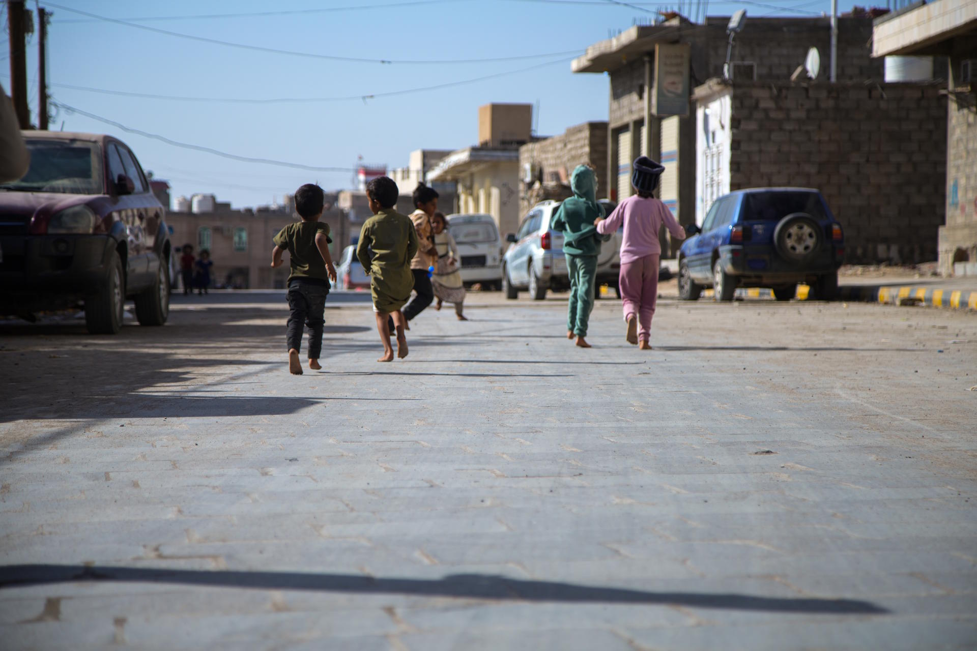 Children running joyfully down a street, with cars parked nearby and buildings in the background.
