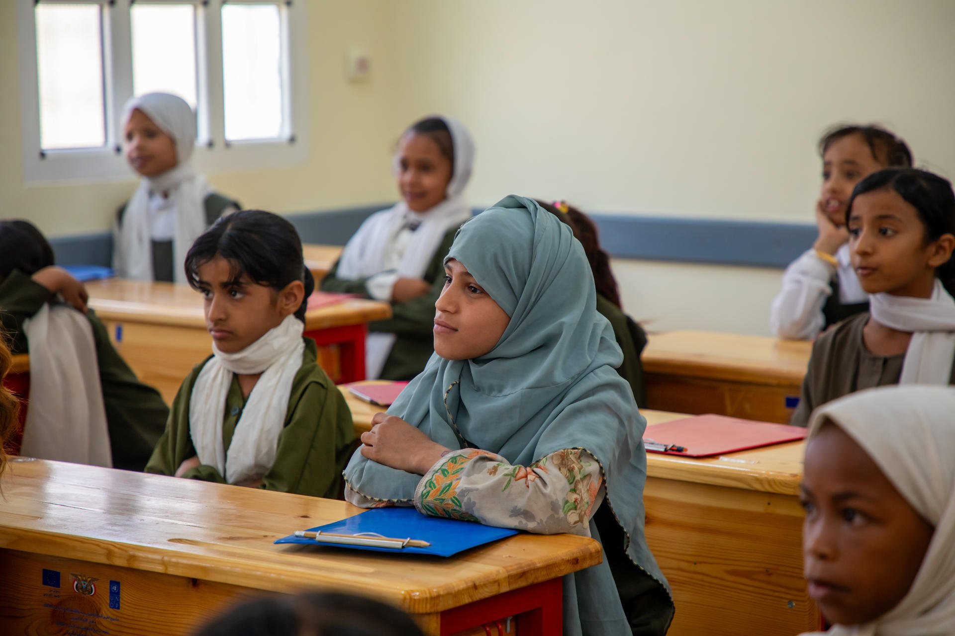 A classroom scene with attentive young girls wearing colorful scarves, seated at wooden desks.