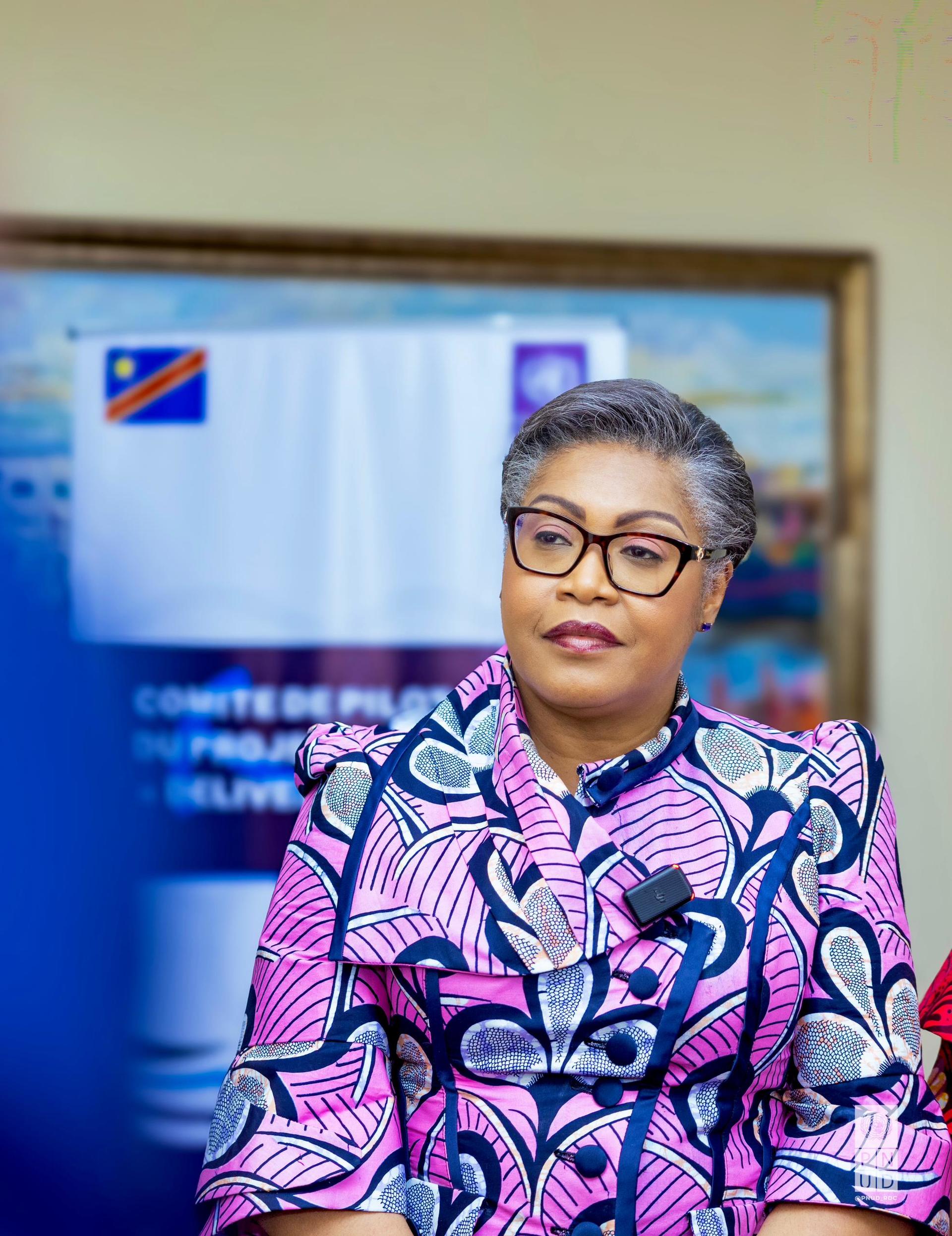 A woman in a colorful patterned blouse sits attentively, facing the camera.