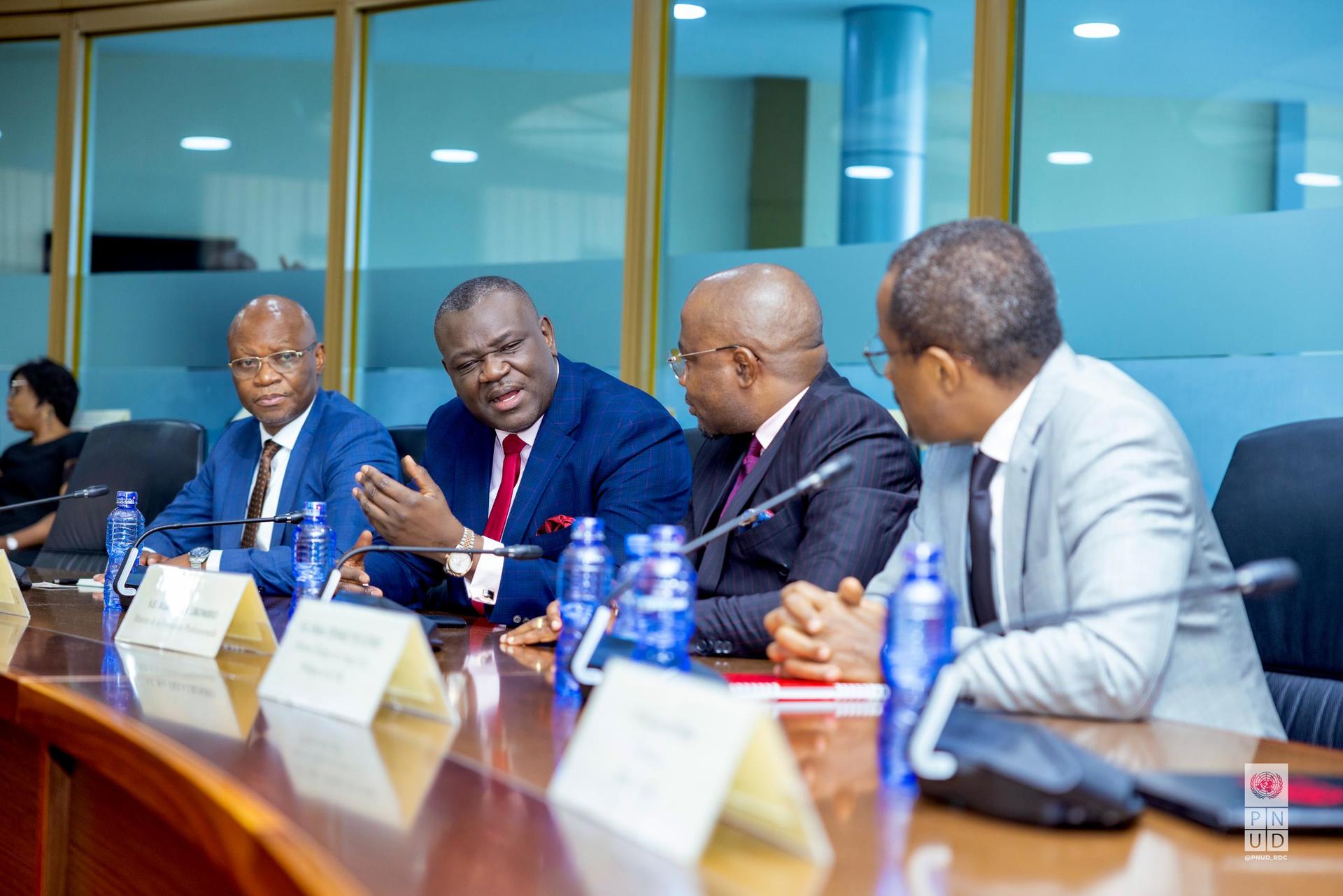 Four men in formal attire seated at a conference table, engaged in conversation.