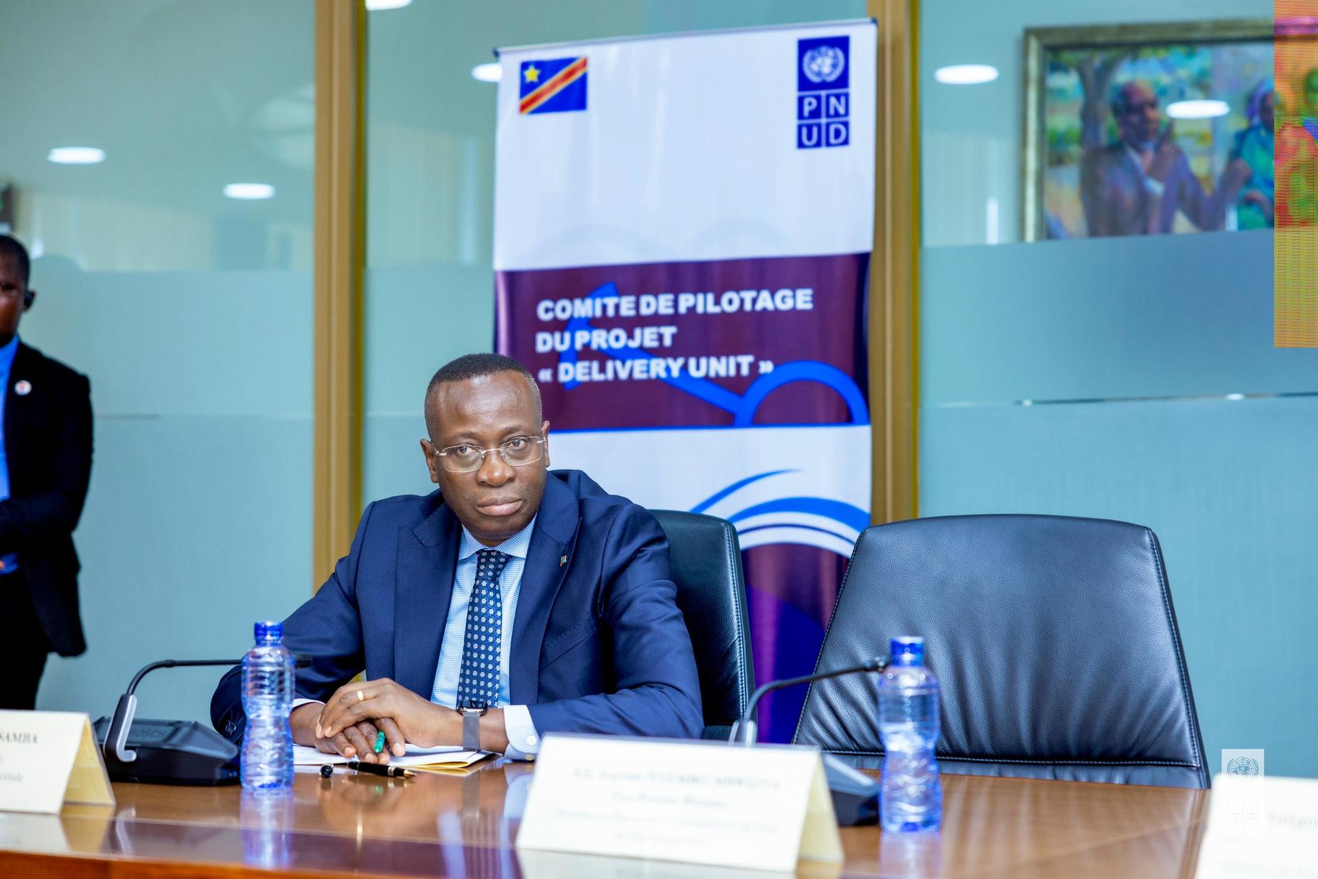 A man in a suit sits at a conference table, looking serious, with banners in the background.