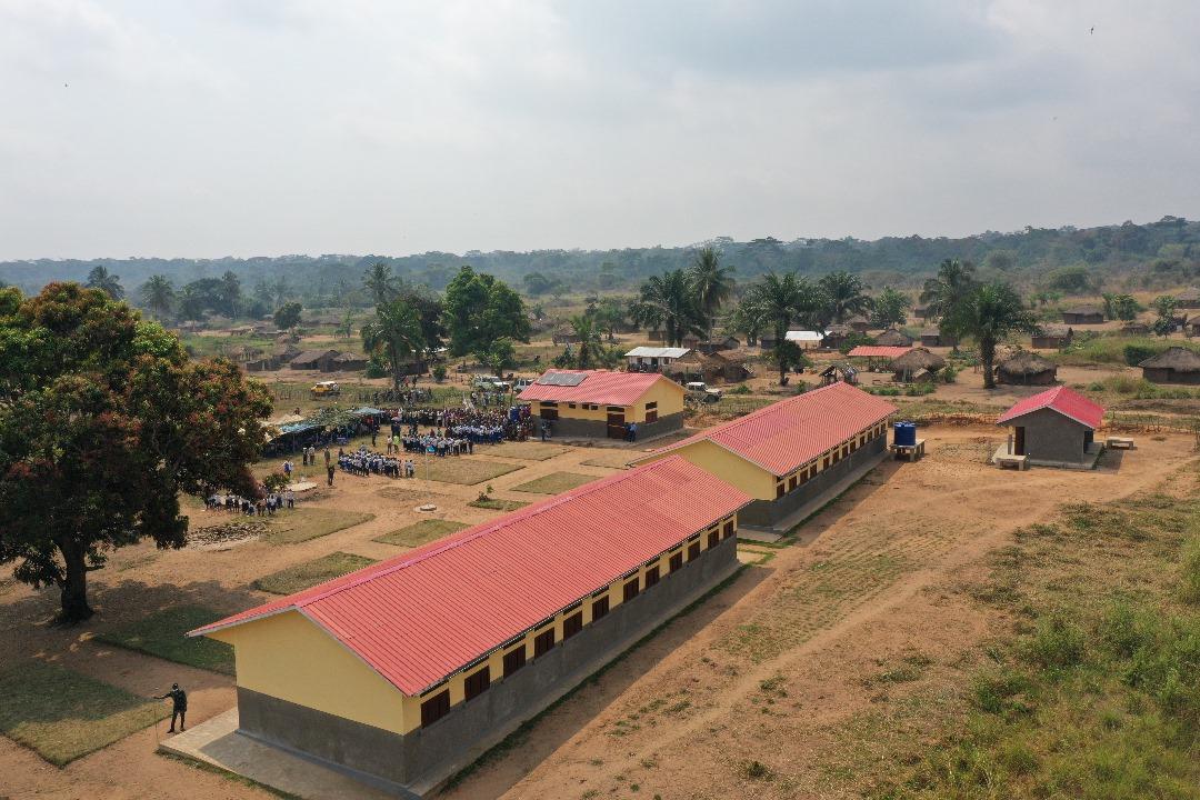 Aerial view of a rural school with red-roofed classrooms amid greenery and dirt paths.