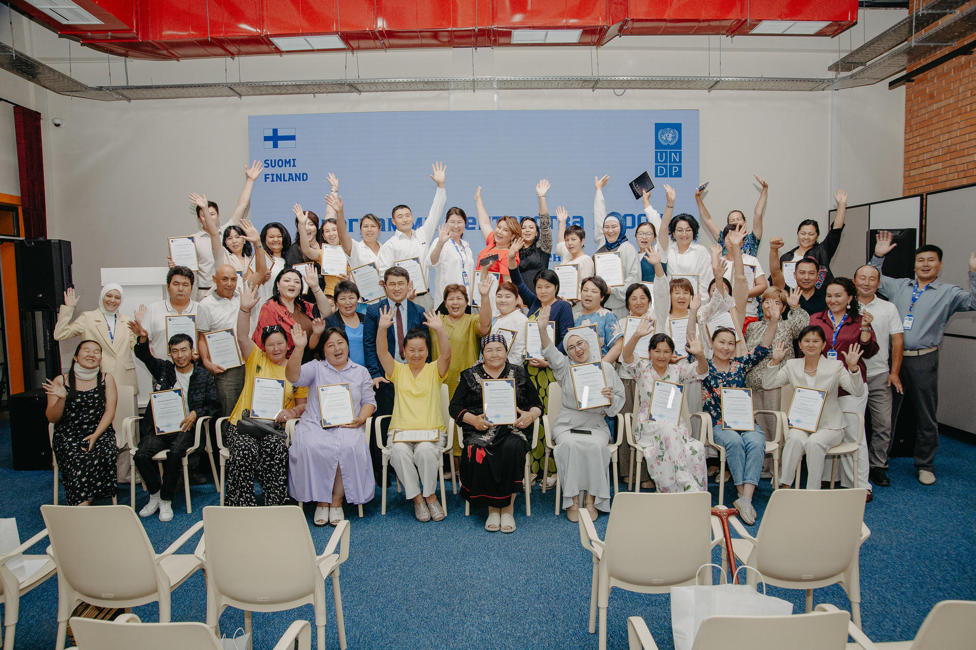 A diverse group of people, seated and standing, smiling and raising their hands in celebration.