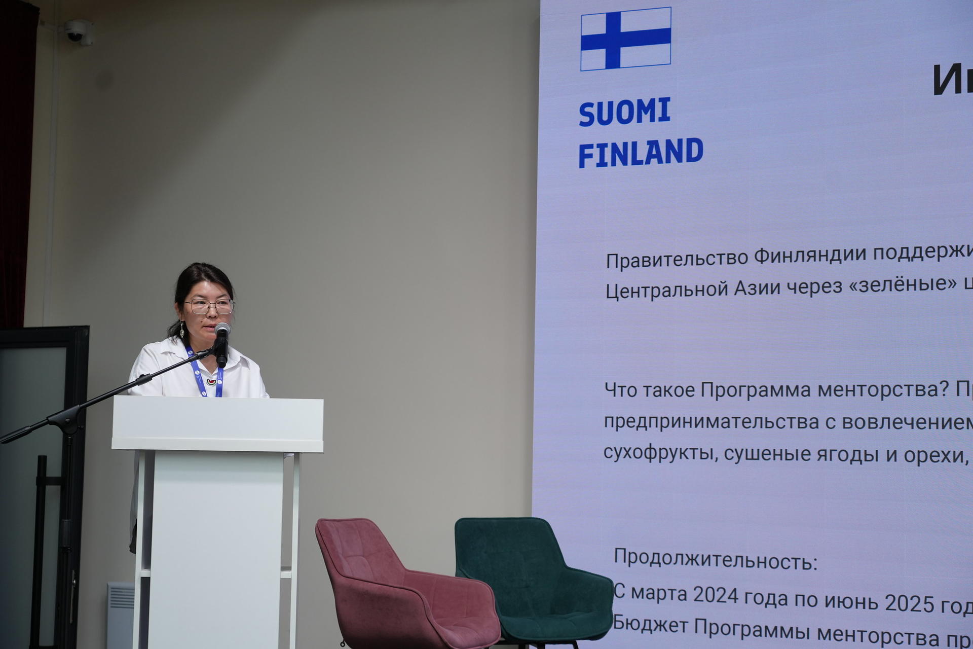 A woman speaks at a podium in front of a presentation screen with Finnish branding.