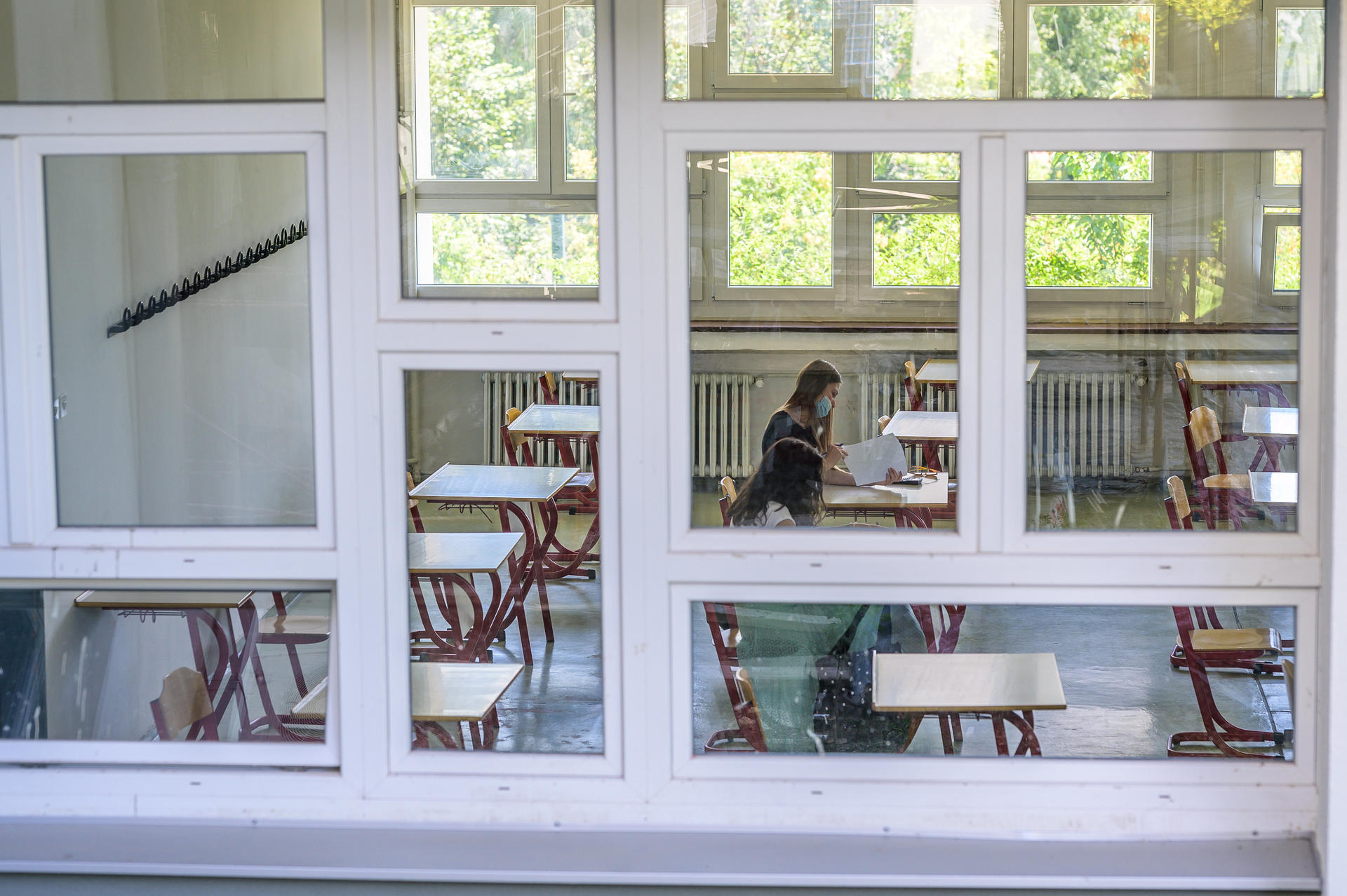 A student sitting at a desk inside a classroom, visible through large windows.