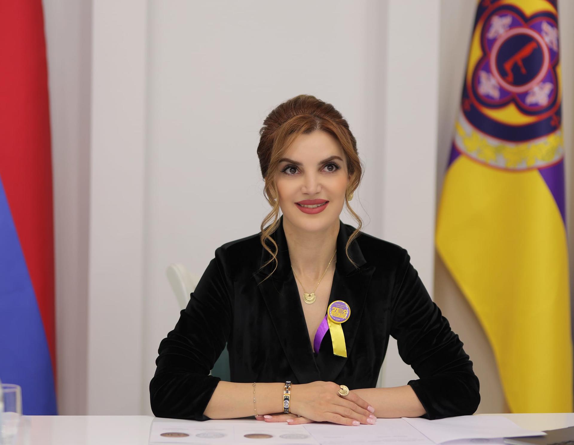 A woman in formal attire sits at a table, smiling, with flags in the background.