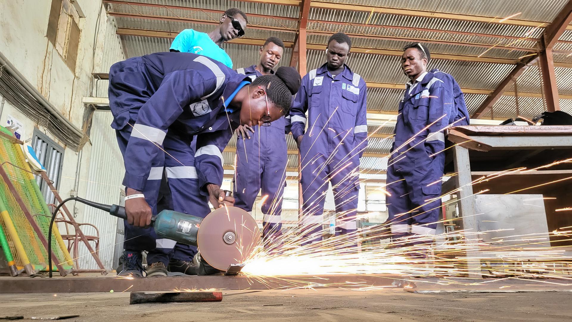 A worker operates a cutting machine, creating sparks, while four observers watch attentively.