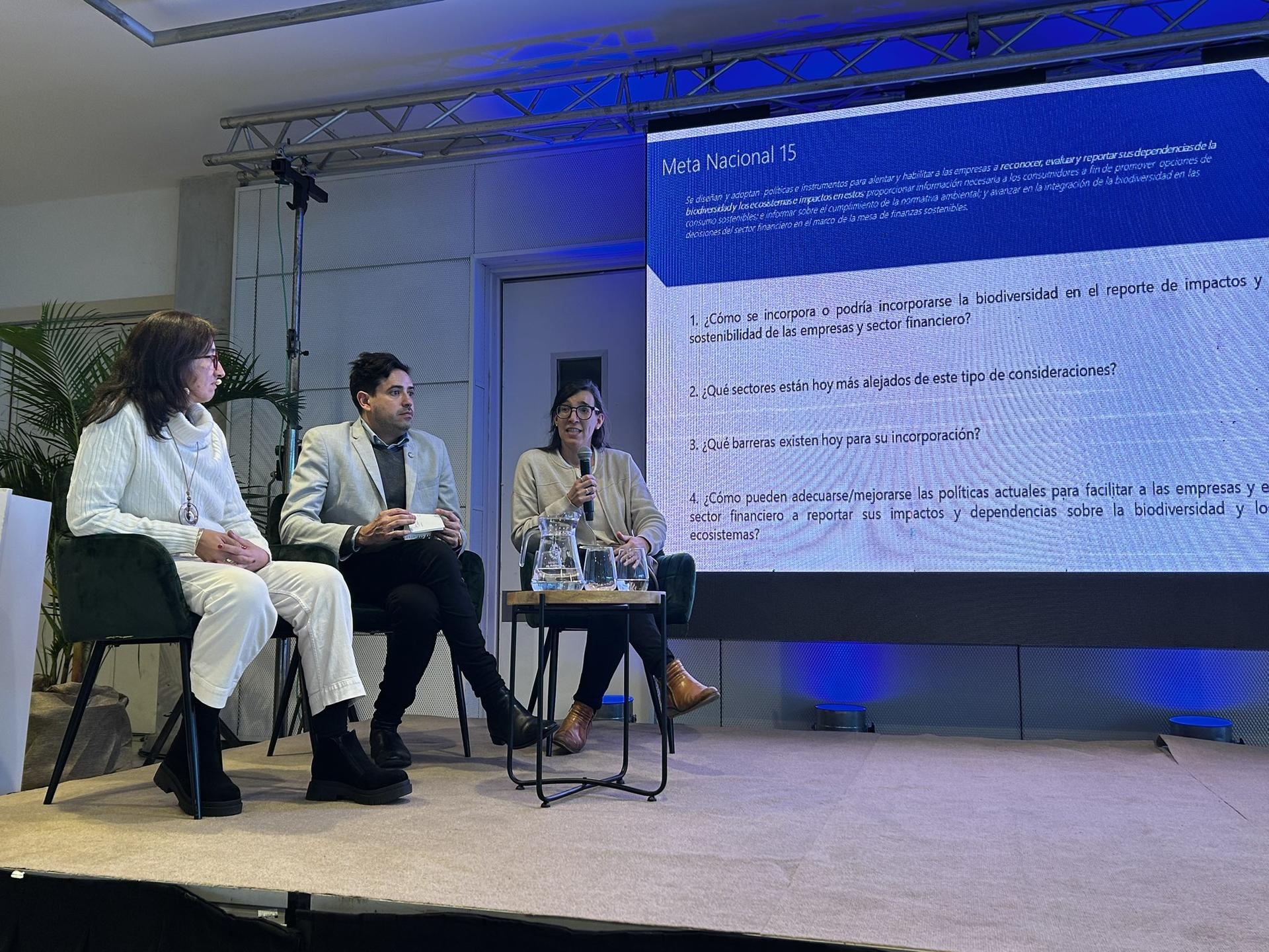 Three panelists seated on stage discussing topics, with a presentation displayed behind them.