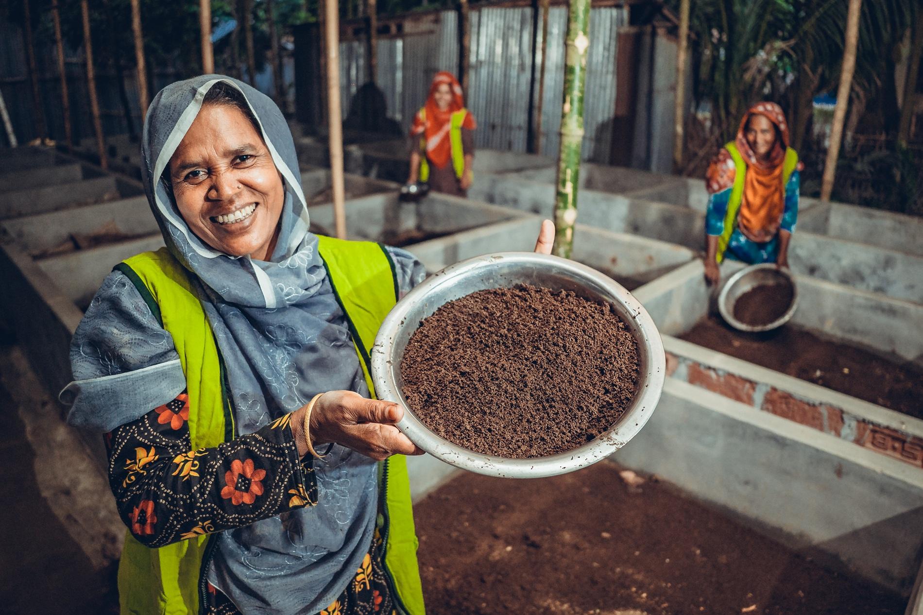 Smiling woman holds a tray of soil in a greenhouse, with other women working in the background.