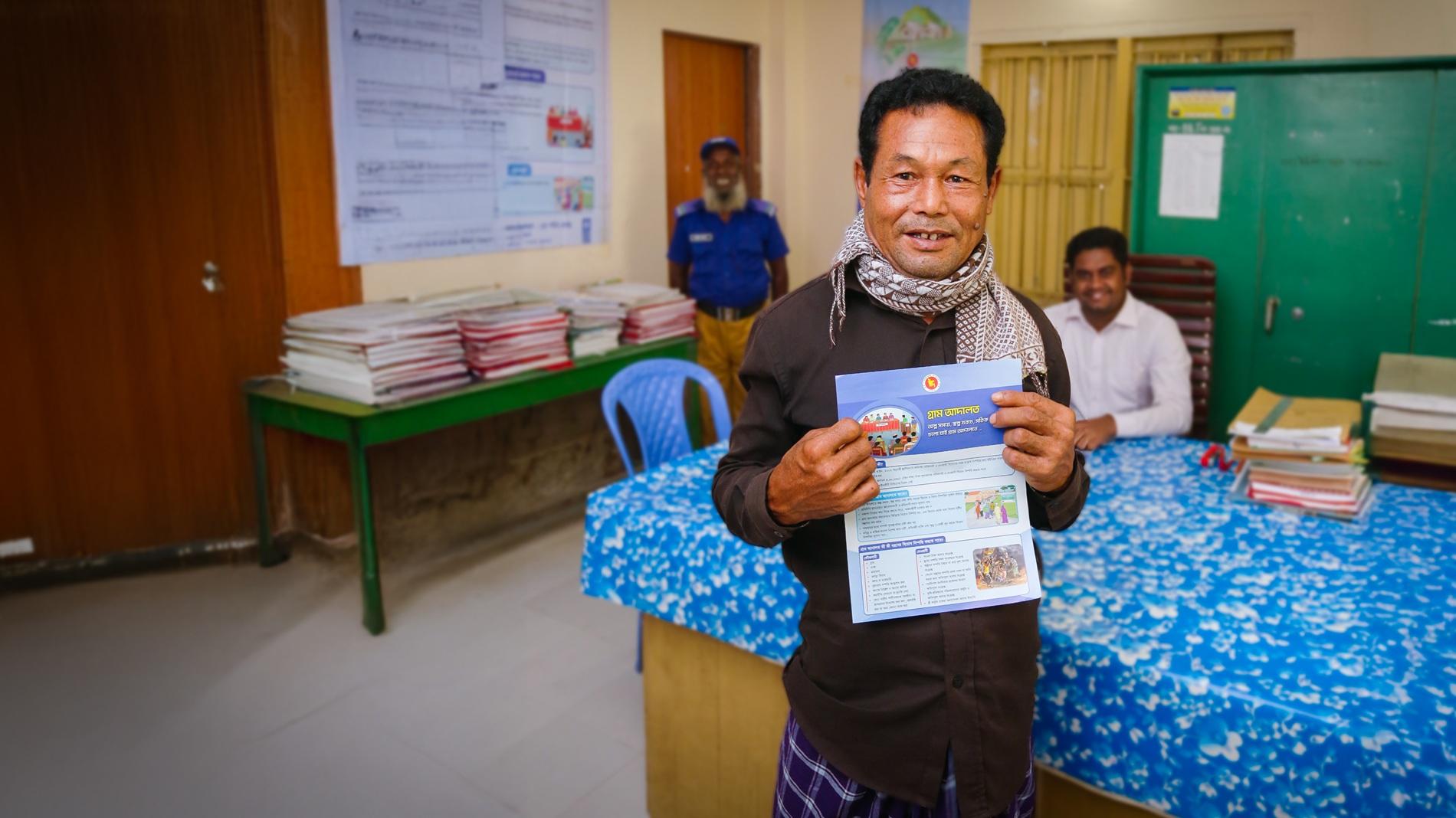 A man holds a document and smiles in an office with a desk and a blue tablecloth.
