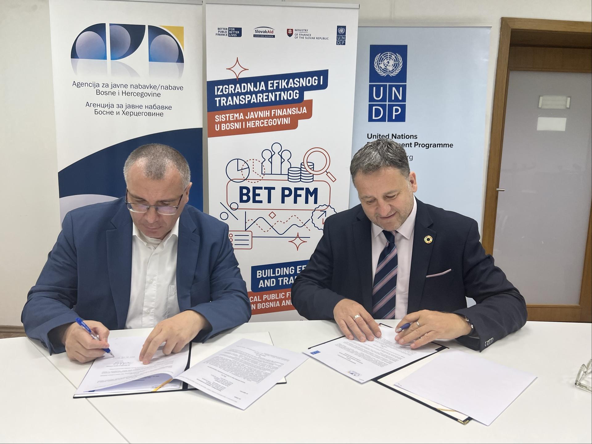 Two men in suits signing documents at a table, with banners in the background.