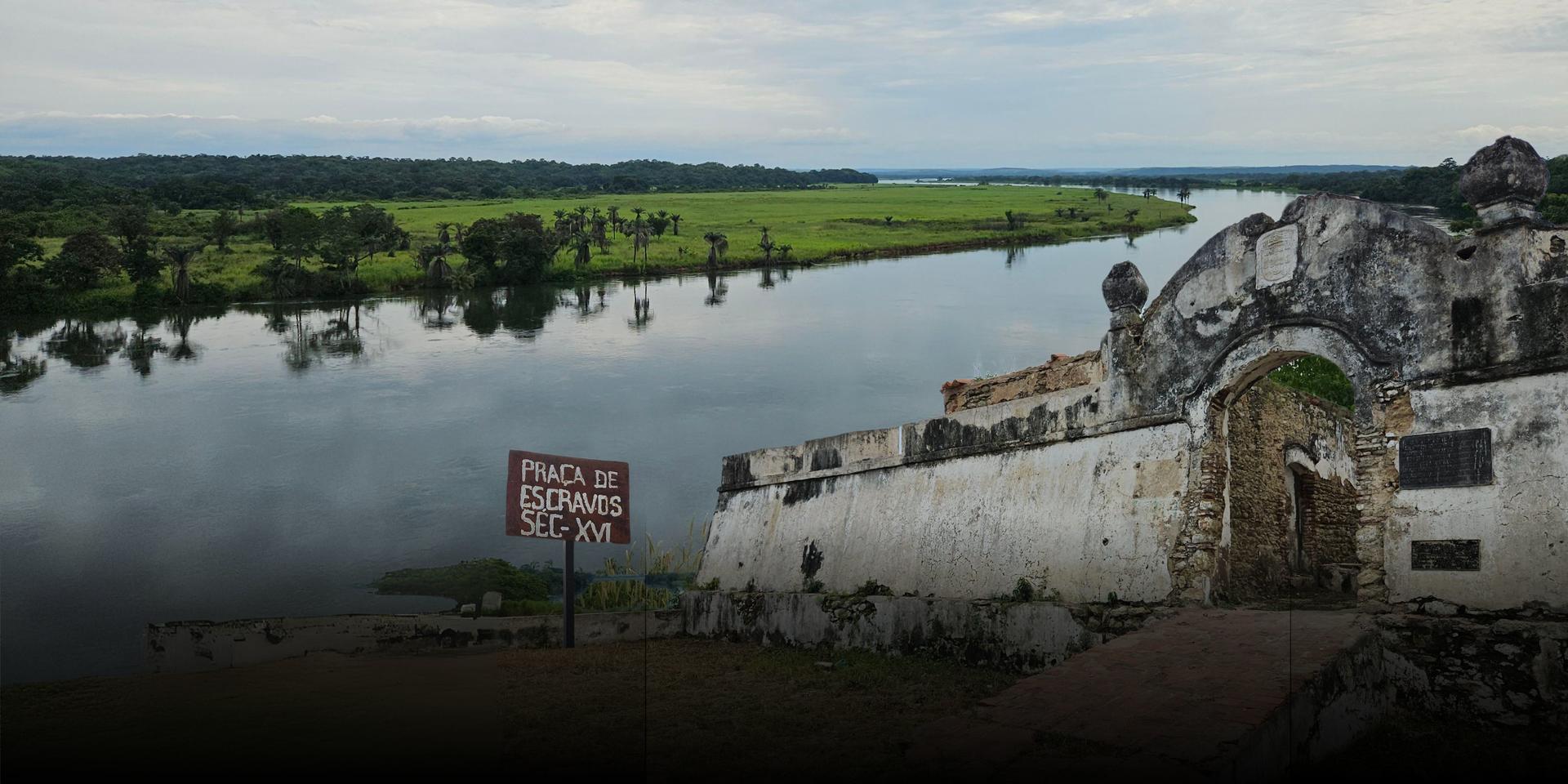 Ancient ruins beside a calm river, lush greenery in the background under a cloudy sky.