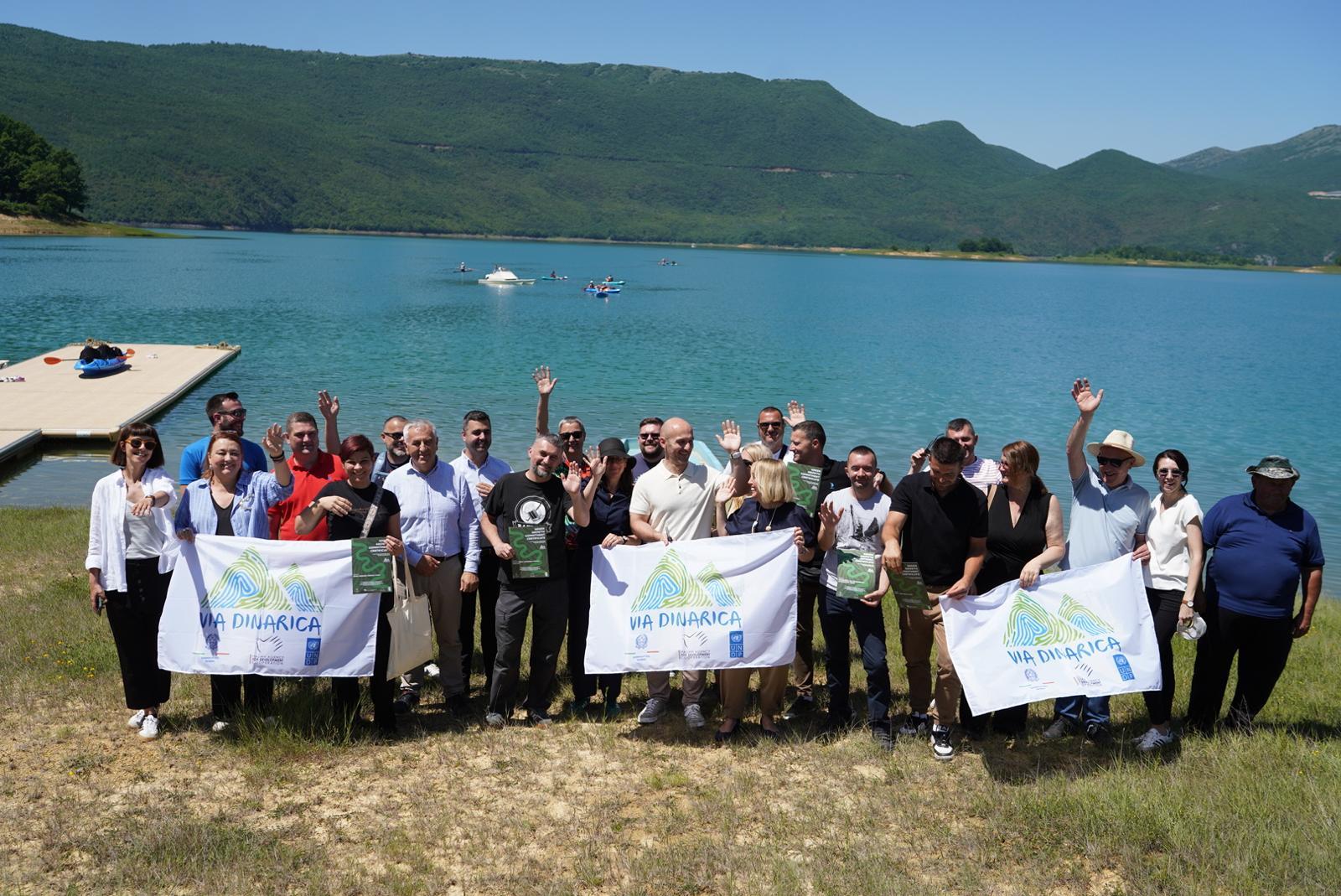 A diverse group of people poses together by a lake, waving and holding banners.