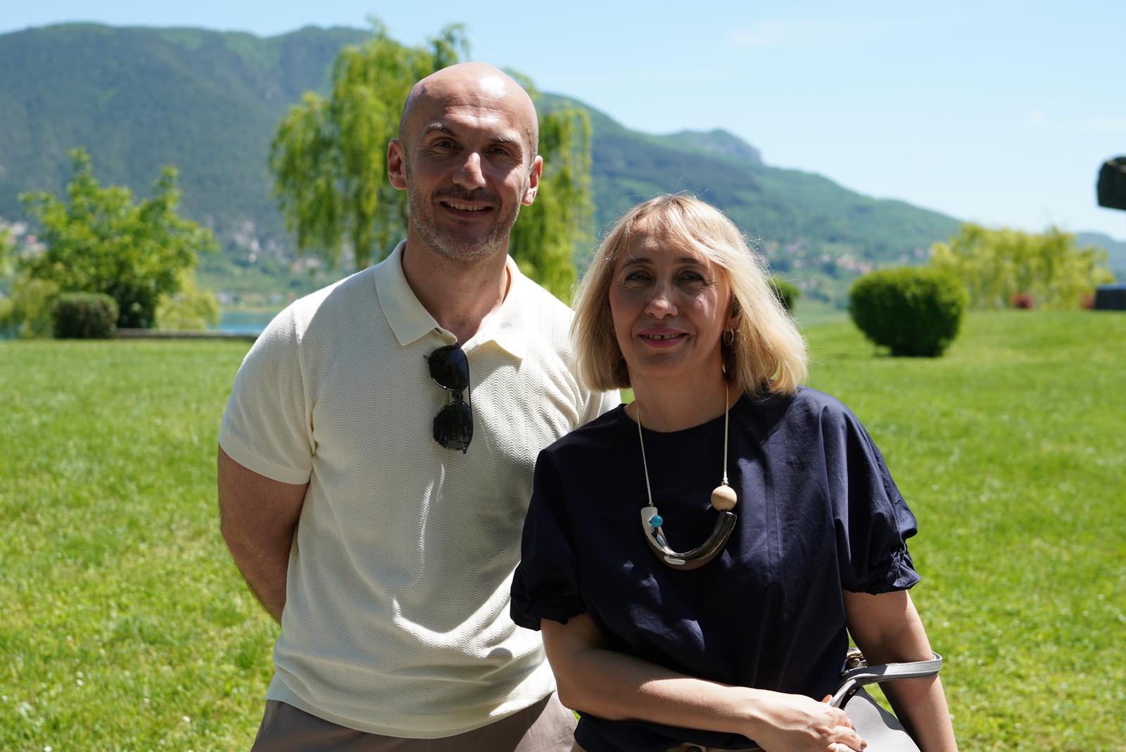 A man and a woman stand together outdoors, smiling, with green hills in the background.