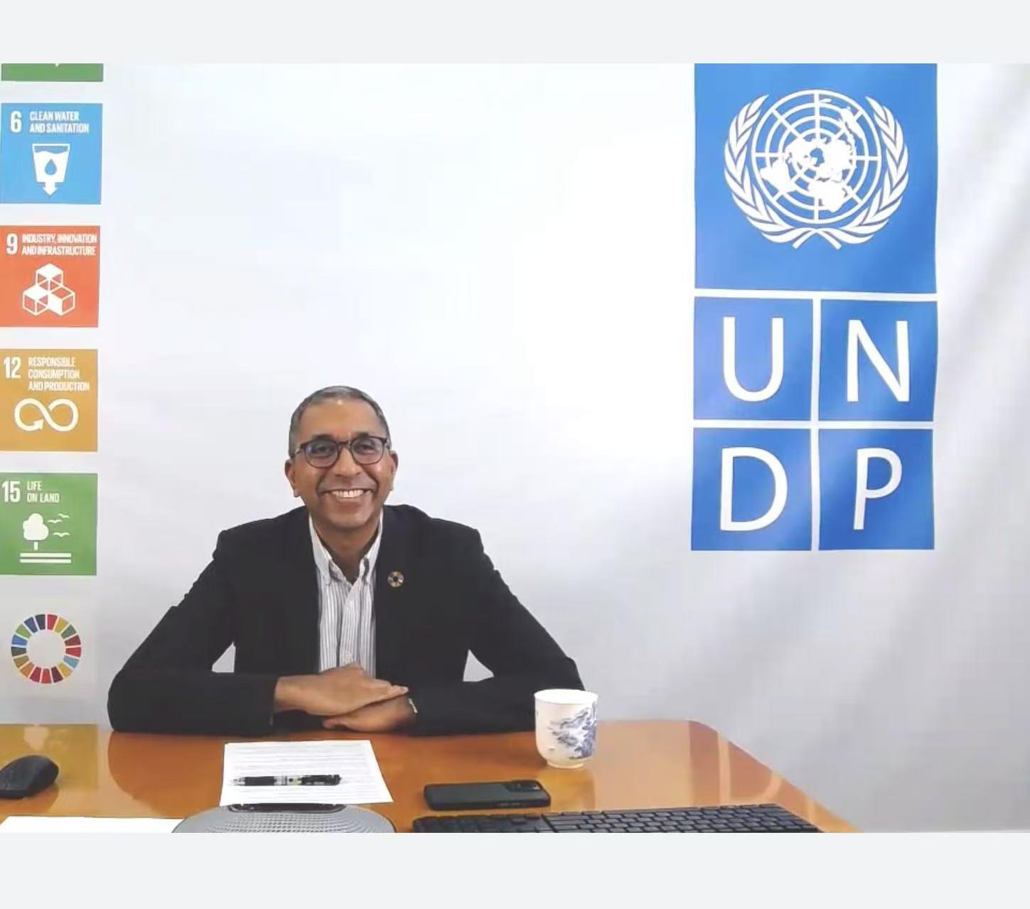 A smiling man in a suit sits at a desk with a UNDP logo and colorful icons in the background.
