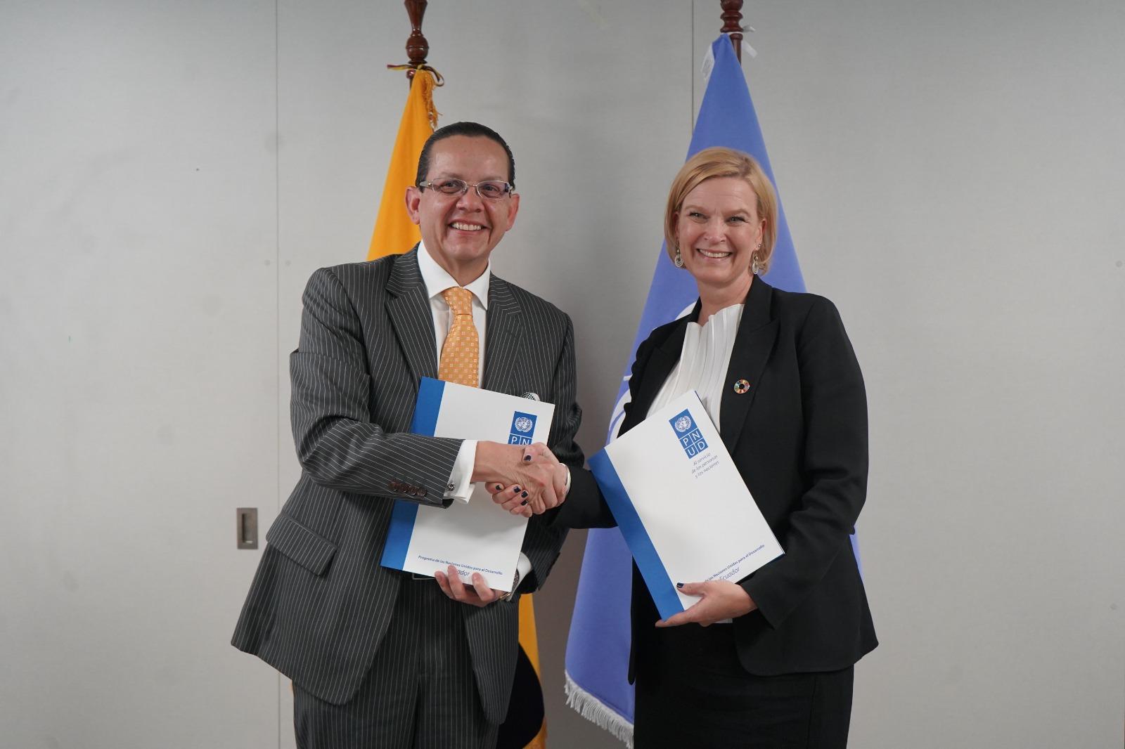 Two individuals in formal attire shaking hands, each holding documents, with flags in the background.