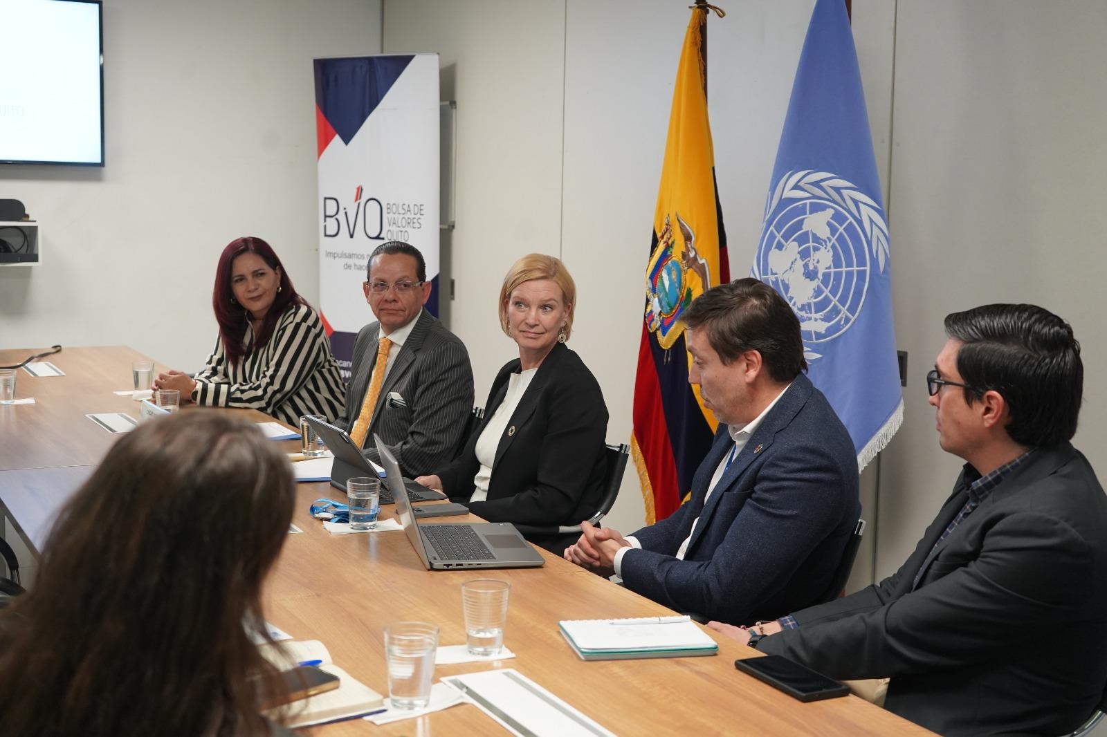 Five professionals sit at a conference table with flags and a projector in the background.