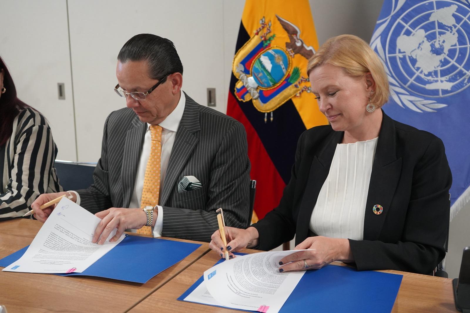 Two officials signing documents at a table, with Ecuador and UN flags in the background.