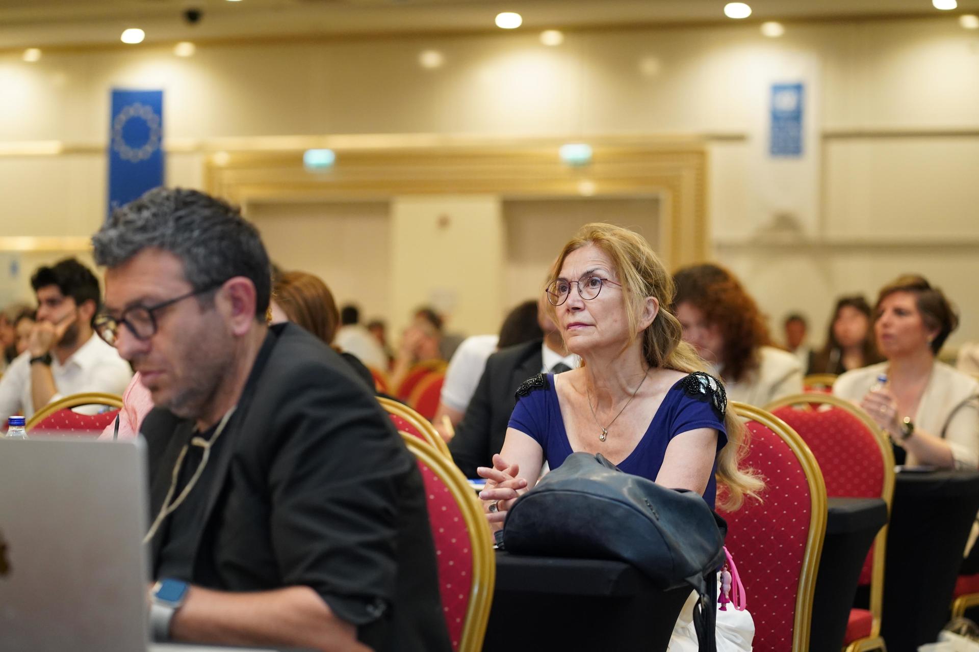 A crowd of people at a conference, with a focus on a woman in a blue dress and a man using a laptop.