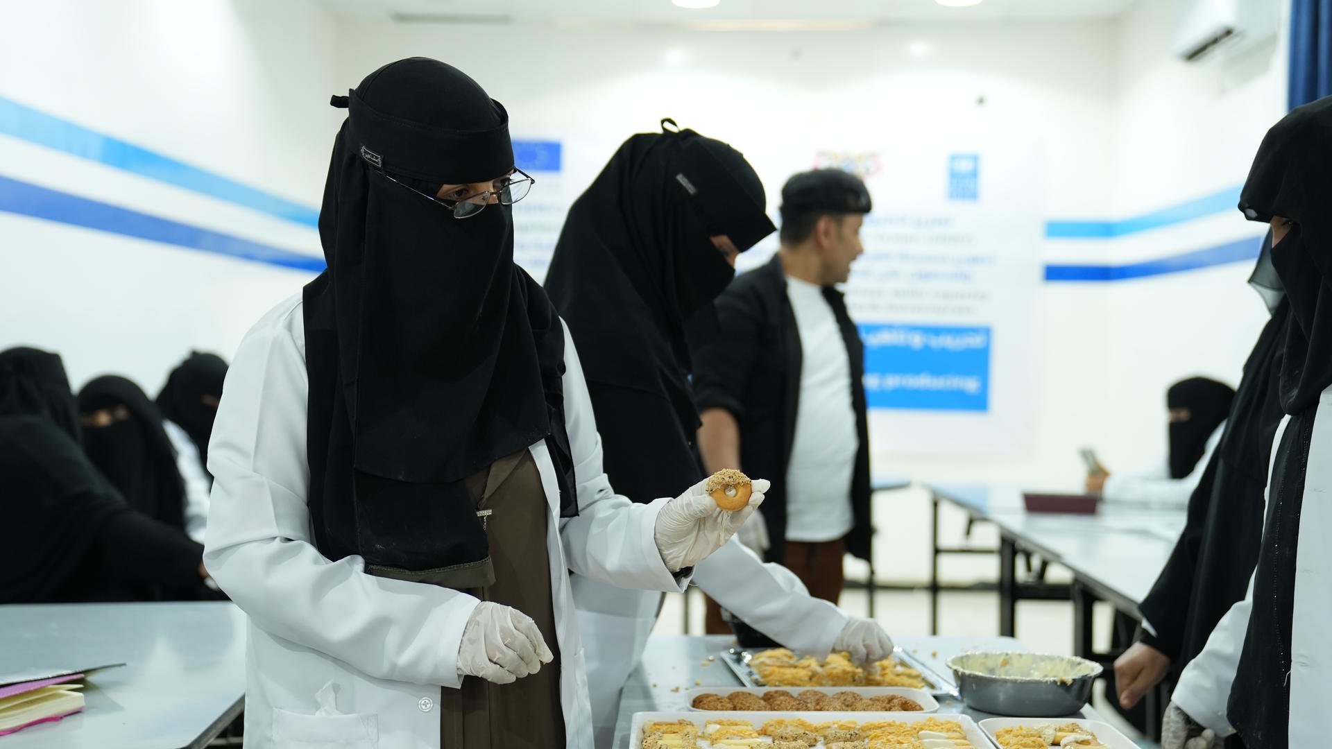 Women in black niqabs and lab coats prepare food in a bright indoor kitchen setting.