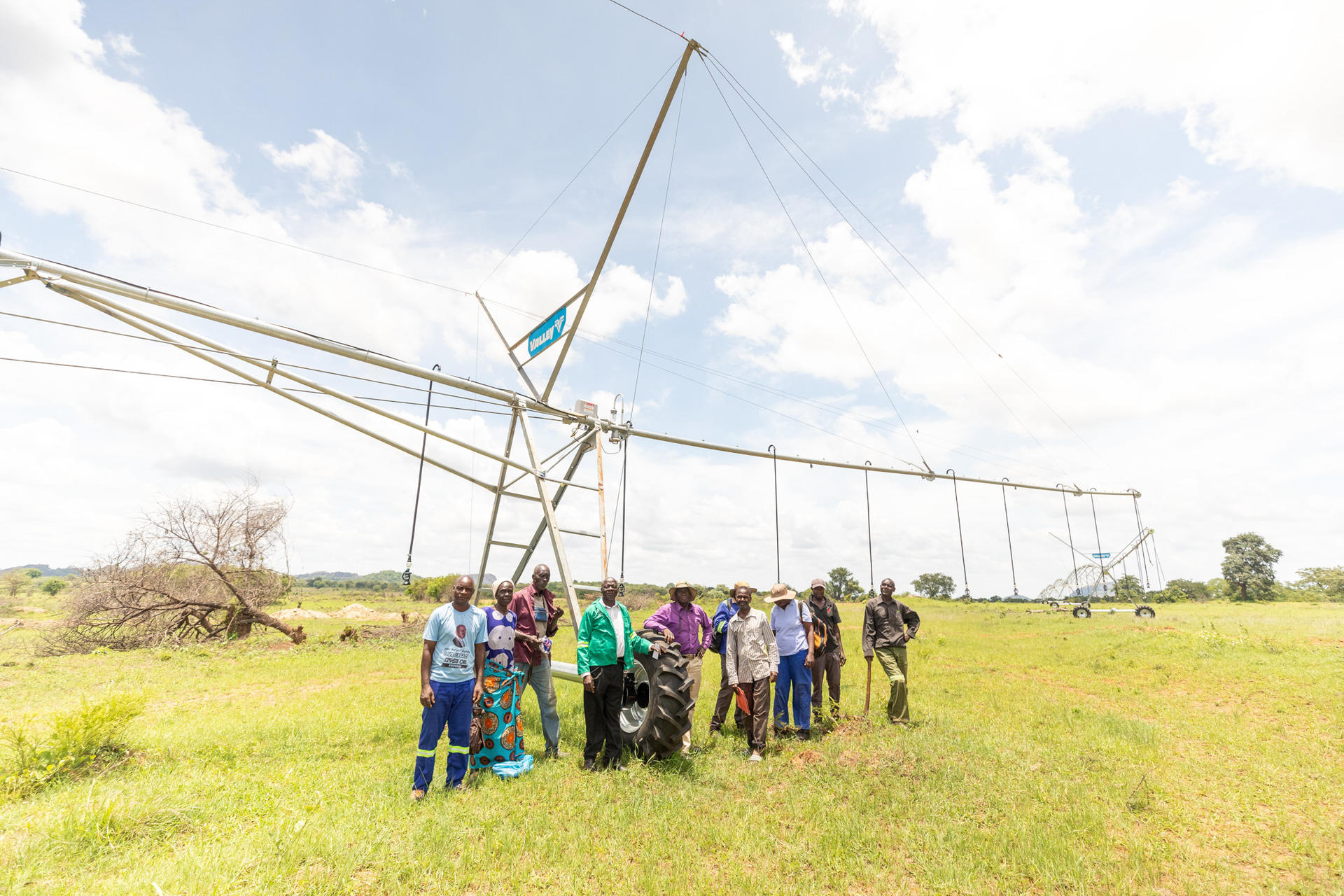 Group of people gathered around a large agricultural structure in a grassy field under a blue sky.