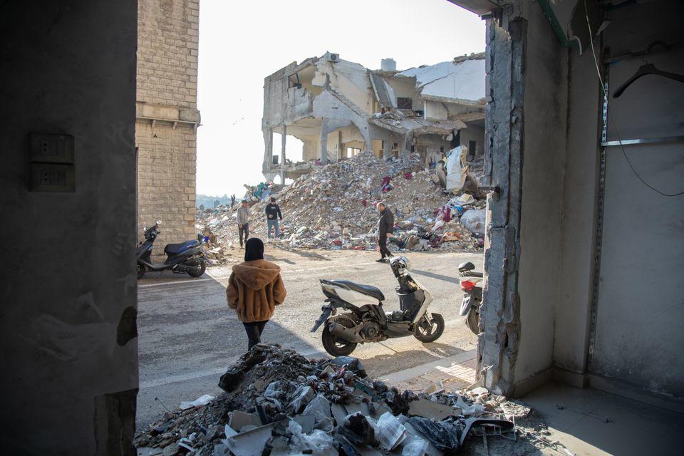 A person walks past a rubble-strewn street, with damaged buildings in the background.