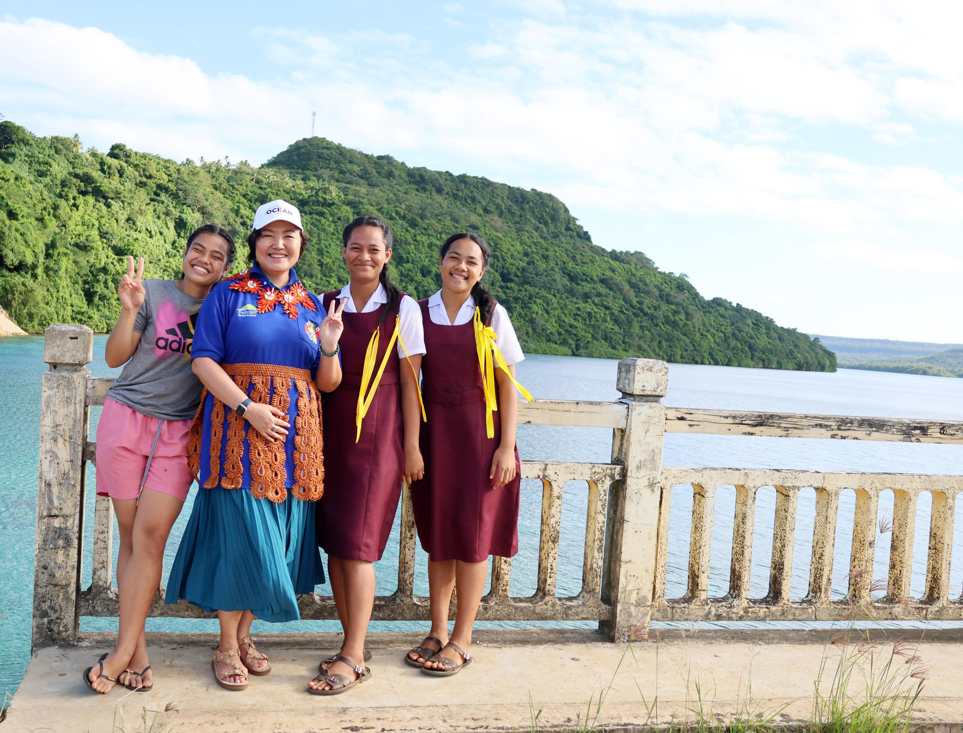 Four smiling young women pose together by a waterfront with lush green hills in the background.