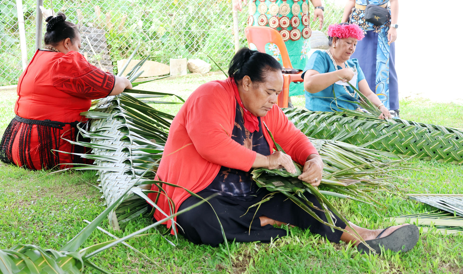 Three women weave with palm leaves on grass, focused on their craft under a canopy.
