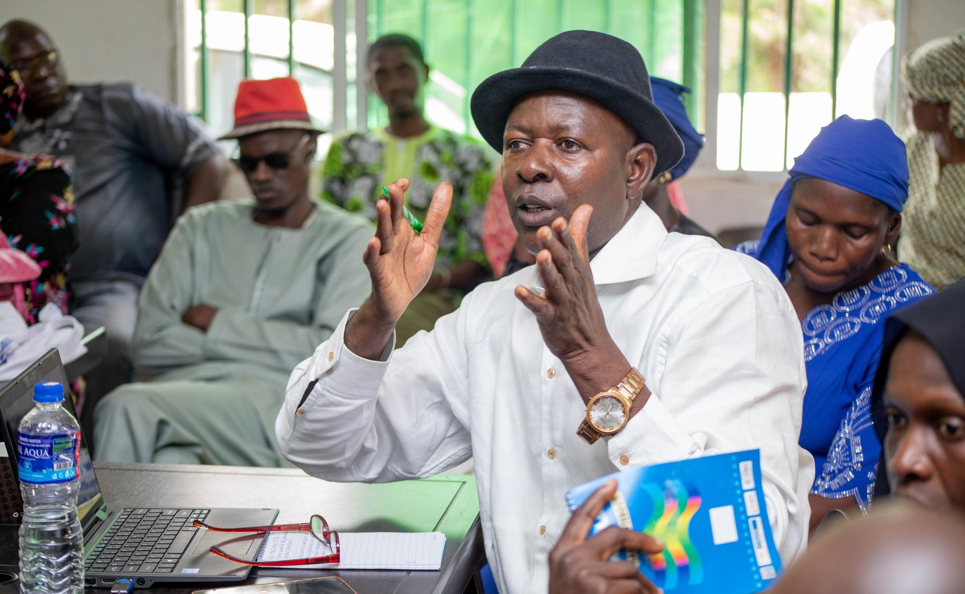 A man in a white shirt and black hat gestures while speaking in a group setting.