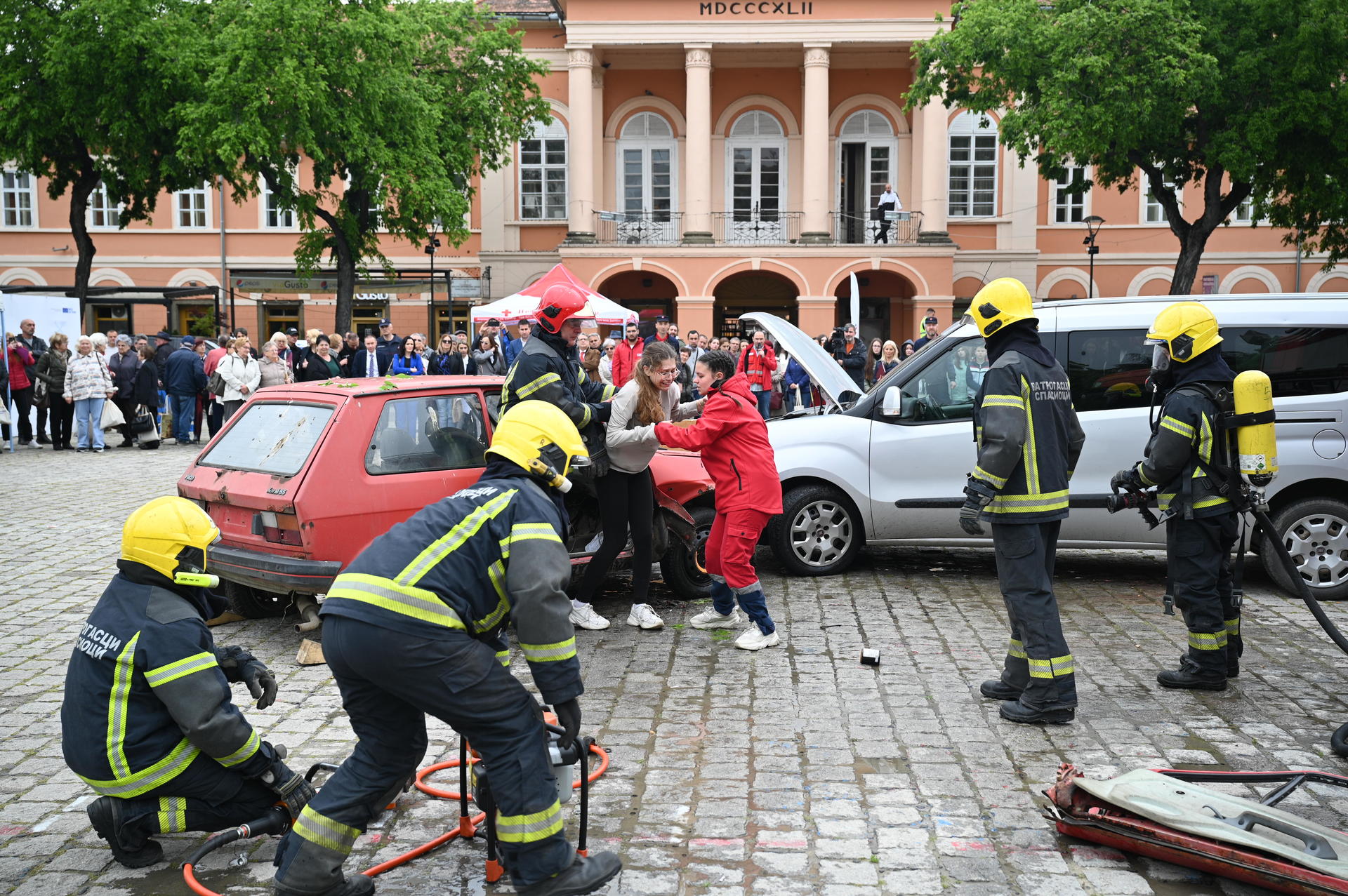 Firefighters respond to an accident scene with a damaged car and bystanders in a square.