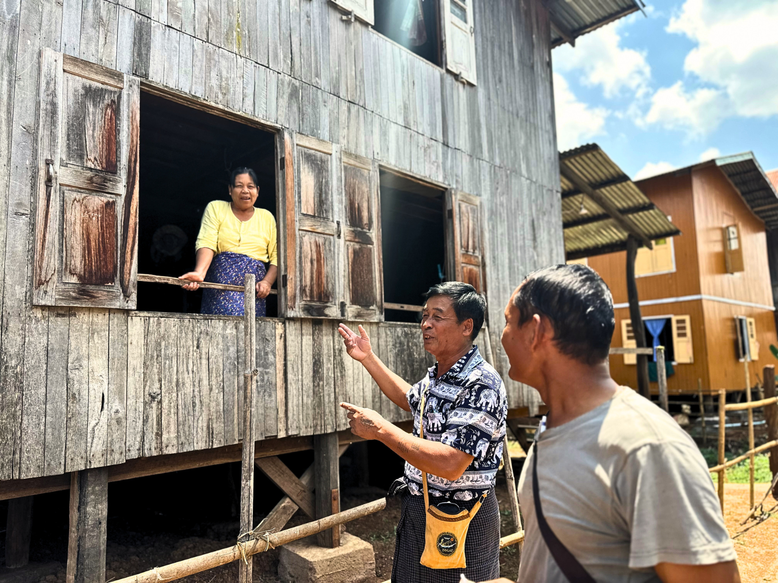 Two men chatting outside a wooden house, while a woman looks out from a window.