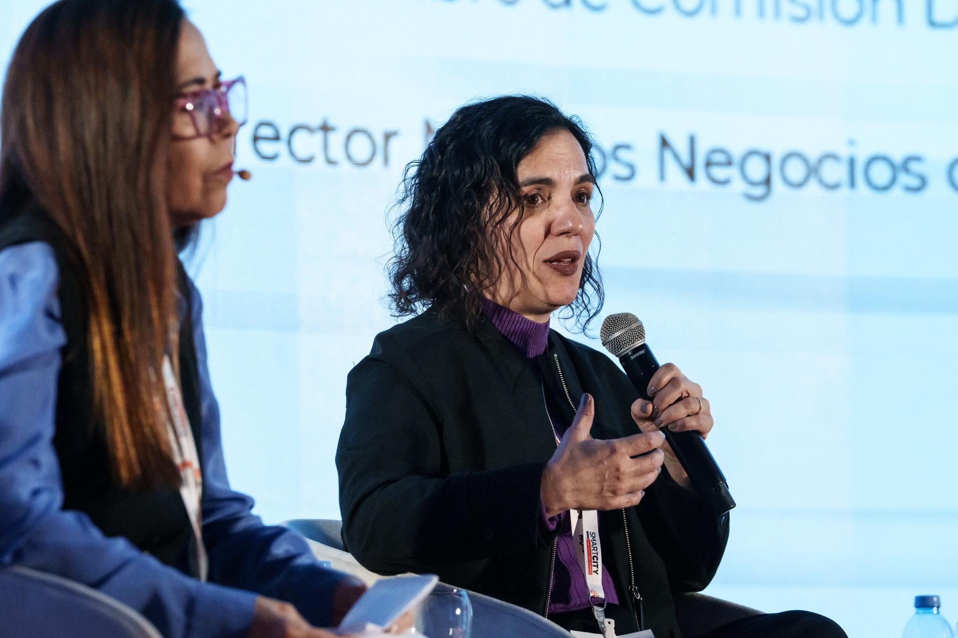 Dos mujeres sentadas en un panel, una hablando con un micrófono.
