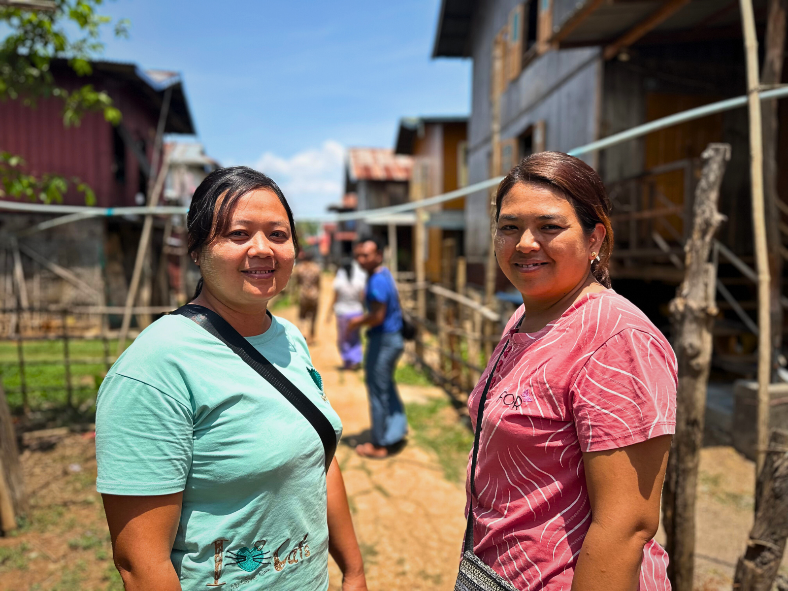 Two young women looking directly at the camera, smiling in a rural setting with wooden houses and a dirt path in the background.