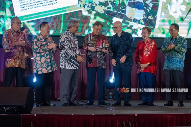 A group of seven individuals in traditional attire gathers on stage, smiling and interacting.