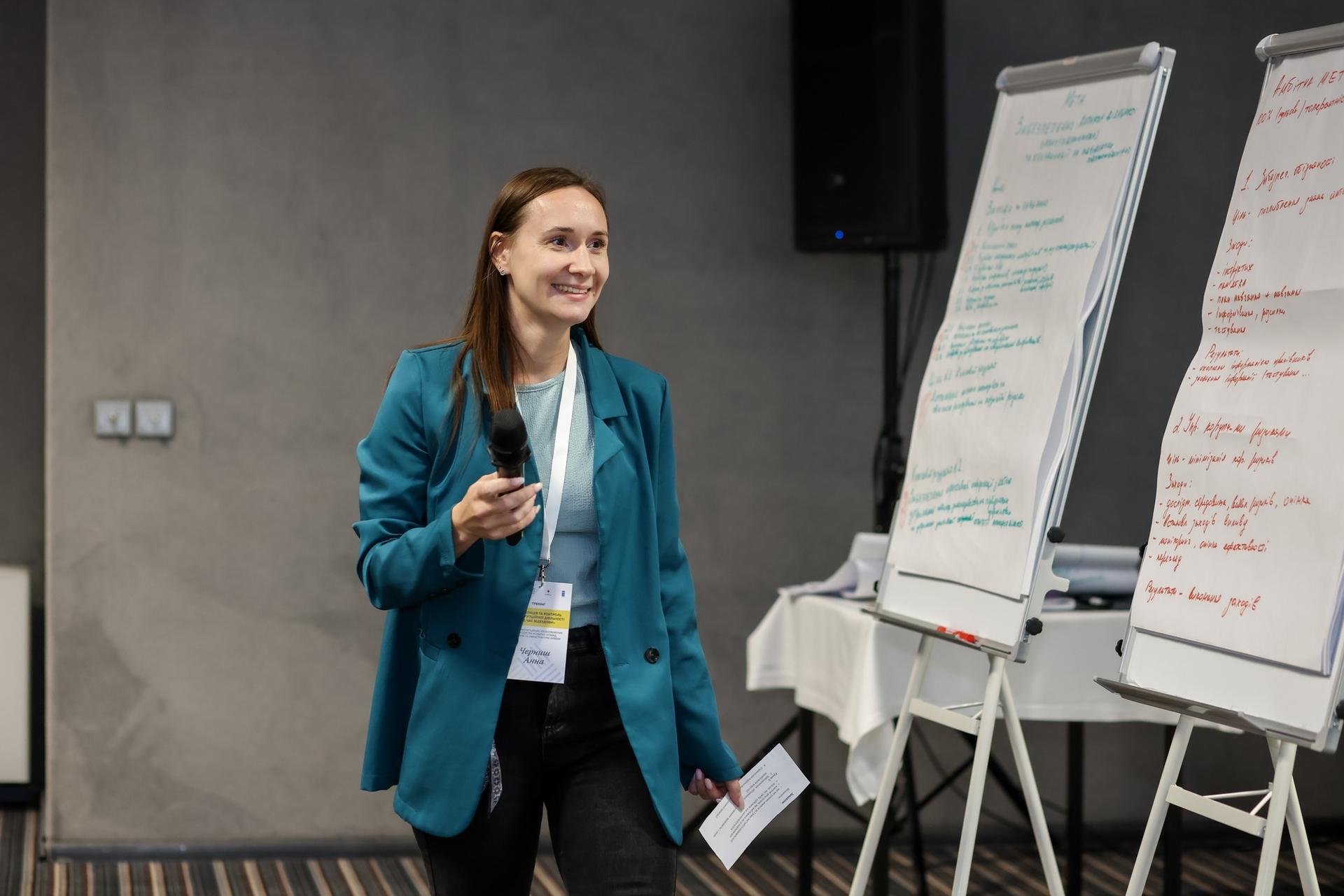 A woman in a teal blazer smiles while holding a microphone, standing near flip charts.