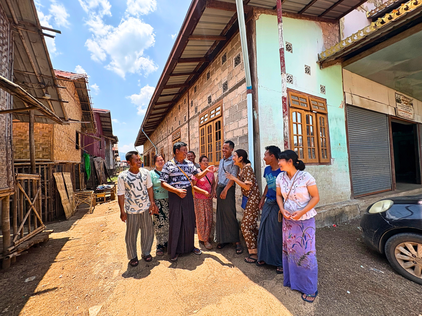 A group of women and men chatting and laughing on a dirt street near traditional houses under a blue sky.