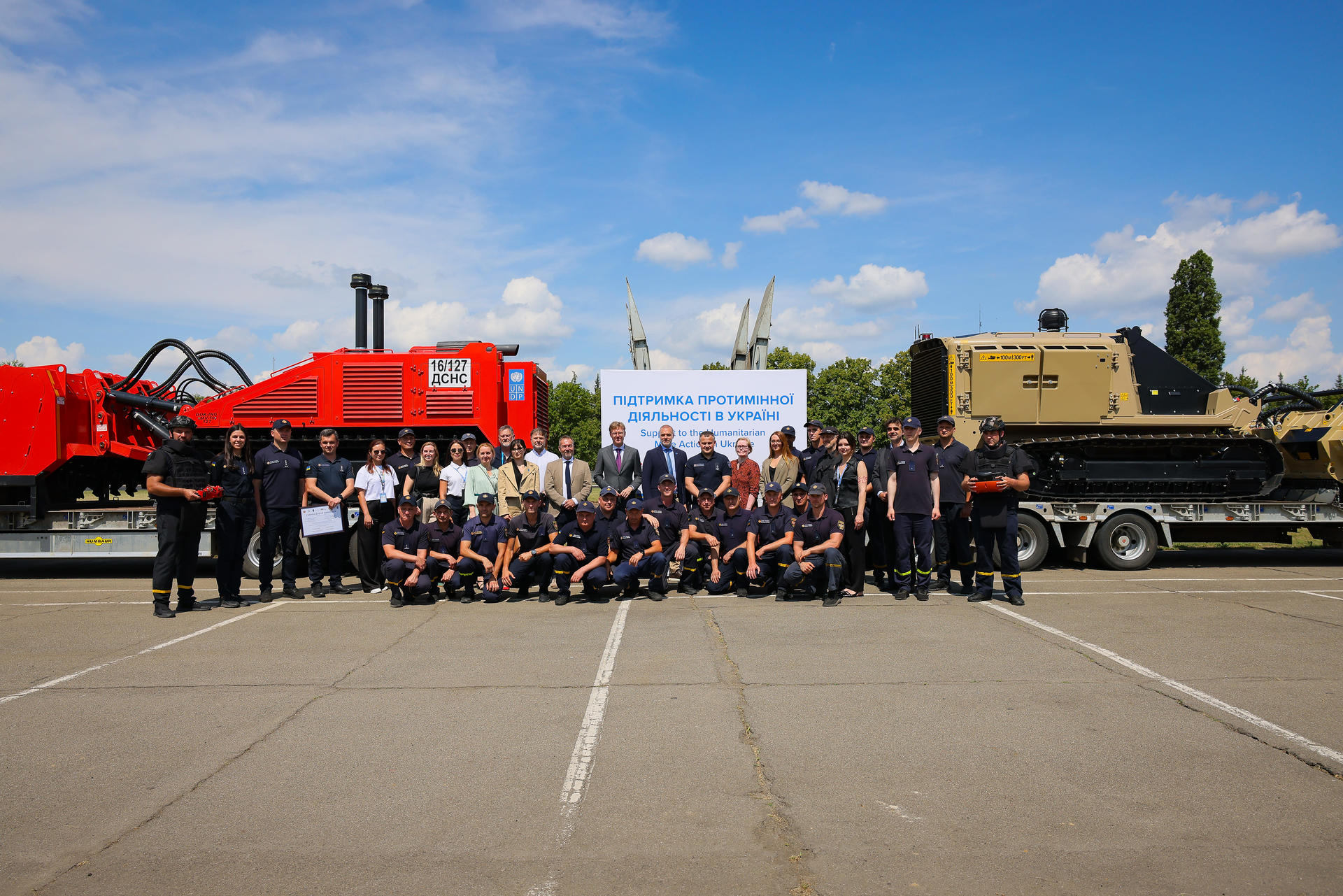 Group photo of people posing with heavy machinery under a blue sky with scattered clouds.