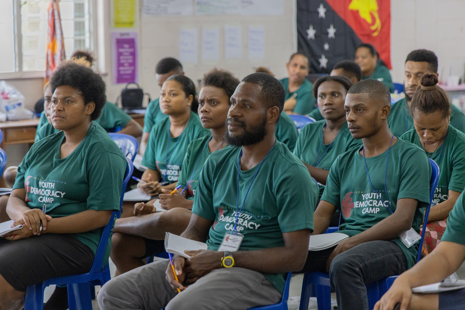 A diverse group of young adults attentively seated in a classroom, wearing green t-shirts.