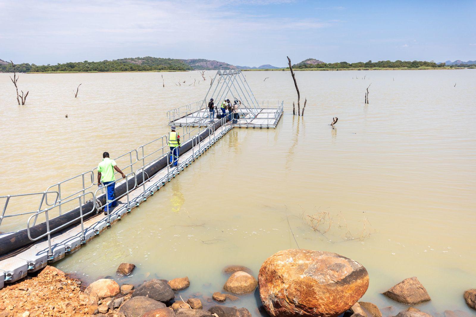 Workers assembling a floating dock on a calm, muddy lake with rocky shores.