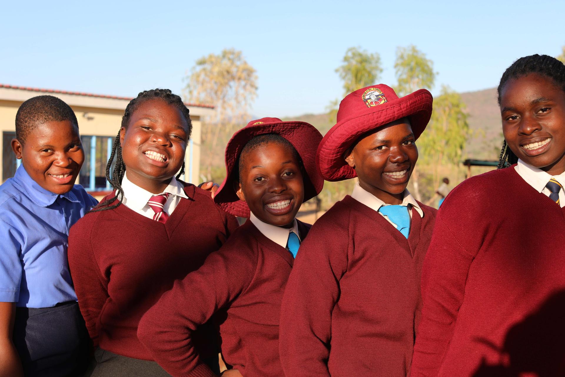 Five smiling schoolgirls in uniforms pose together outdoors, wearing maroon sweaters and hats.
