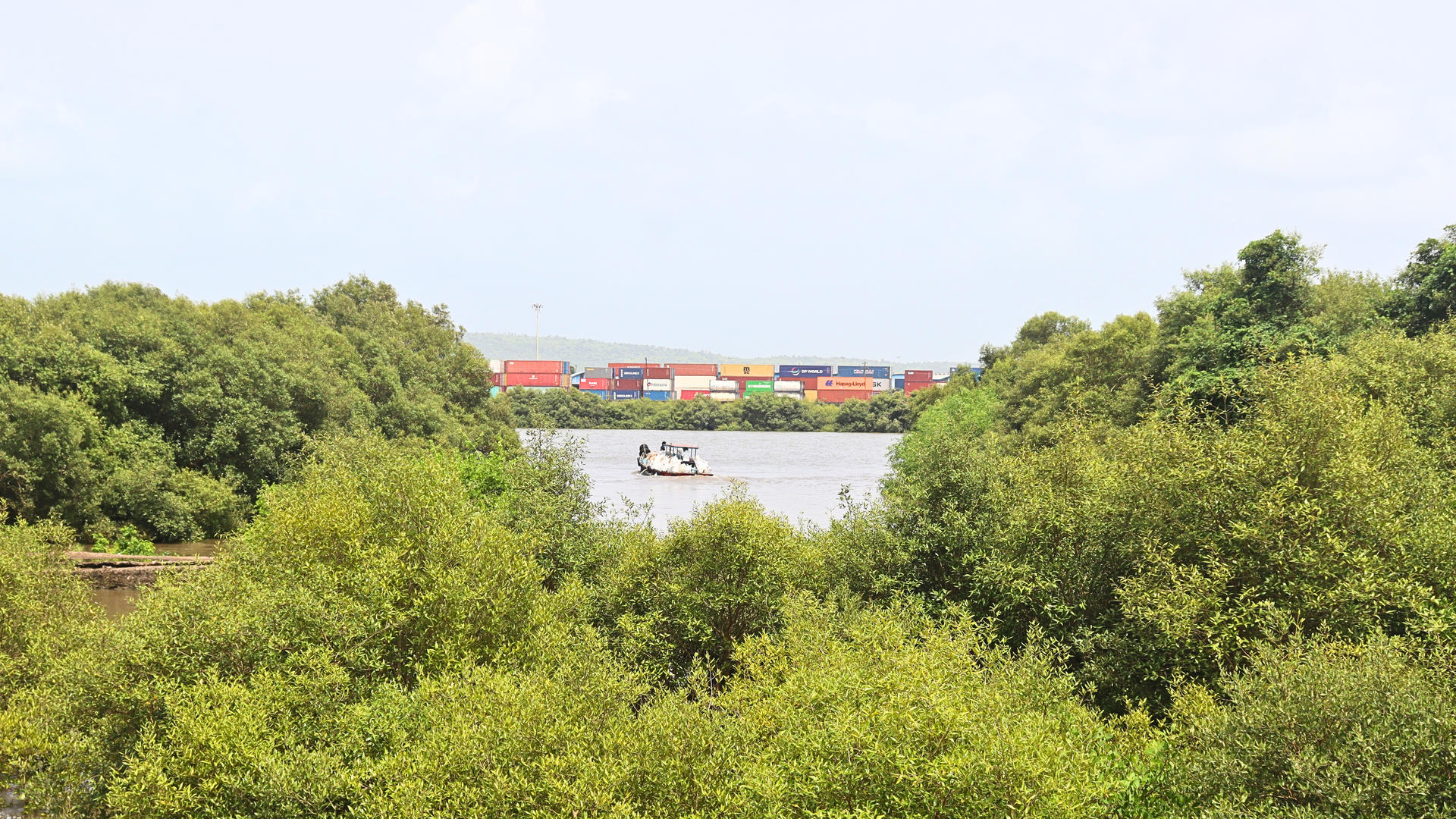 Lush green trees frame a river with a small boat and distant shipping containers.
