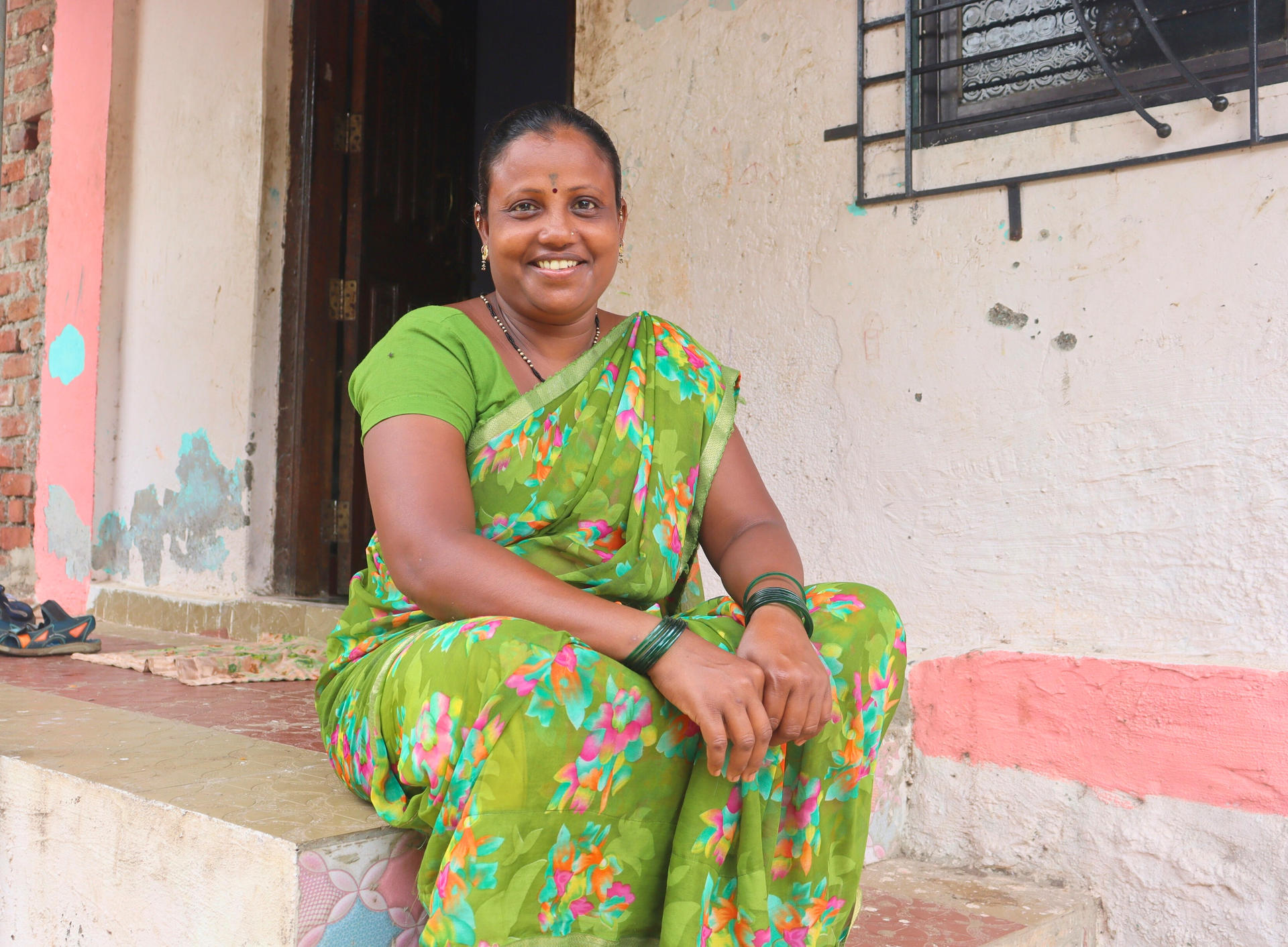 Smiling woman in a green floral saree sitting on steps of a house.