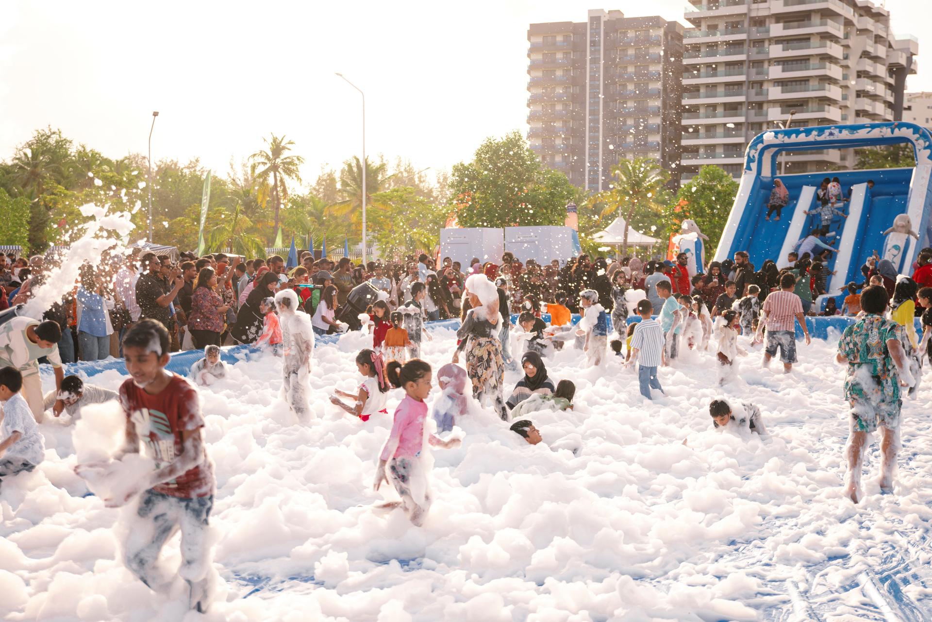 Children playing in a foam-filled area, with a slide and crowd in the background.